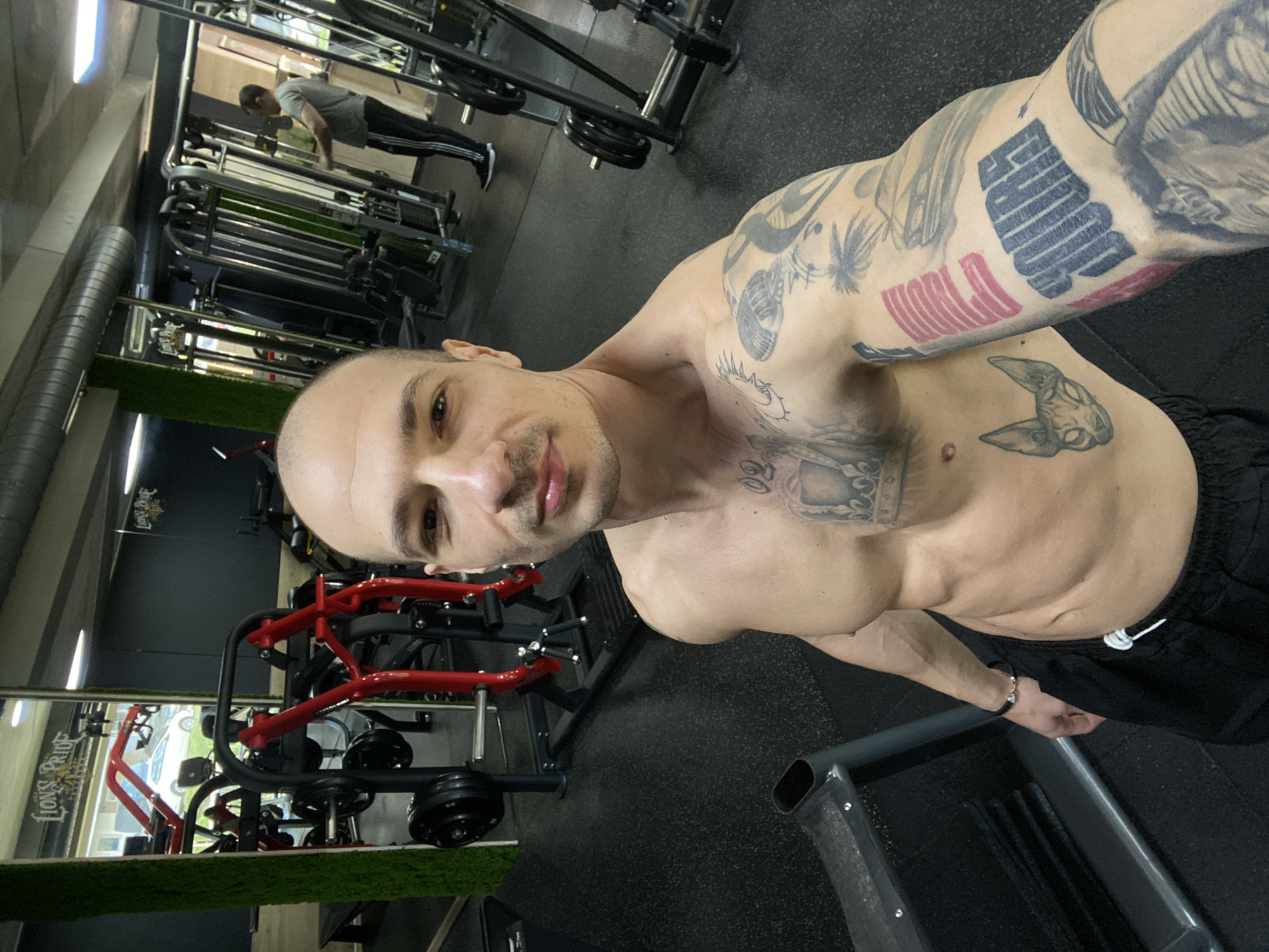 A shirtless man with tattoos is in a gym. The gym has weight machines and the name "LIONS PRIDE" is visible. Another man is in the background, working out. The location is in Pleven, Bulgaria.