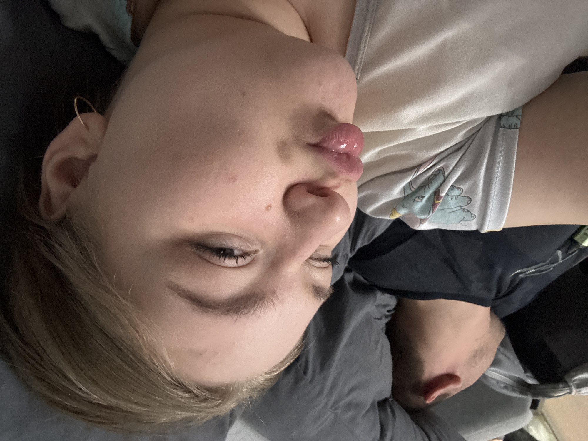 A close-up, angled shot depicts two people lying down. A woman with light skin, blonde hair, and light eyes is in the foreground. She is wearing a light-colored top with a cartoon print on the shoulder. Her lips are pursed. In the background, a man with fair skin and short dark hair is visible. He is wearing a dark-colored shirt. The background appears to be a gray-toned bed or couch. The location is not evident in the image and cannot be determined to be Chaikovsky, Russia.