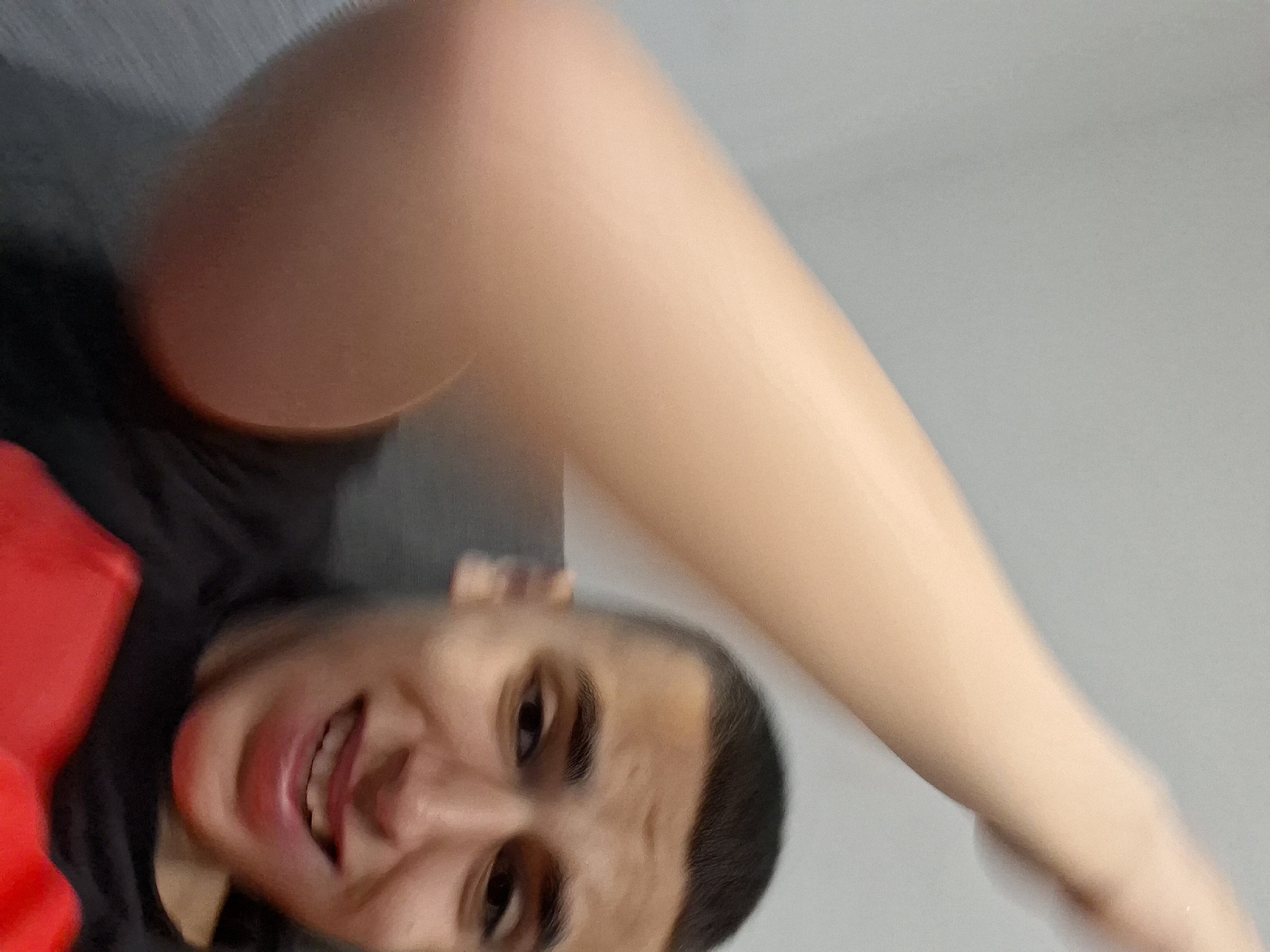 A close-up, slightly blurred image features a young male subject with short, dark hair. His face, positioned in the lower right quadrant, exhibits a wide smile with visible upper teeth and dark eyes looking towards the viewer's right. He wears a dark-colored (likely black) crew-neck shirt, with a prominent red section visible across the lower chest, possibly an undershirt or part of a graphic. His right arm, heavily motion-blurred and light-skinned, extends from the lower-left to the upper-left of the frame, suggesting an upward or sweeping motion. The background is an undifferentiated light grey wall, appearing out of focus. The overall scene indicates an indoor setting.