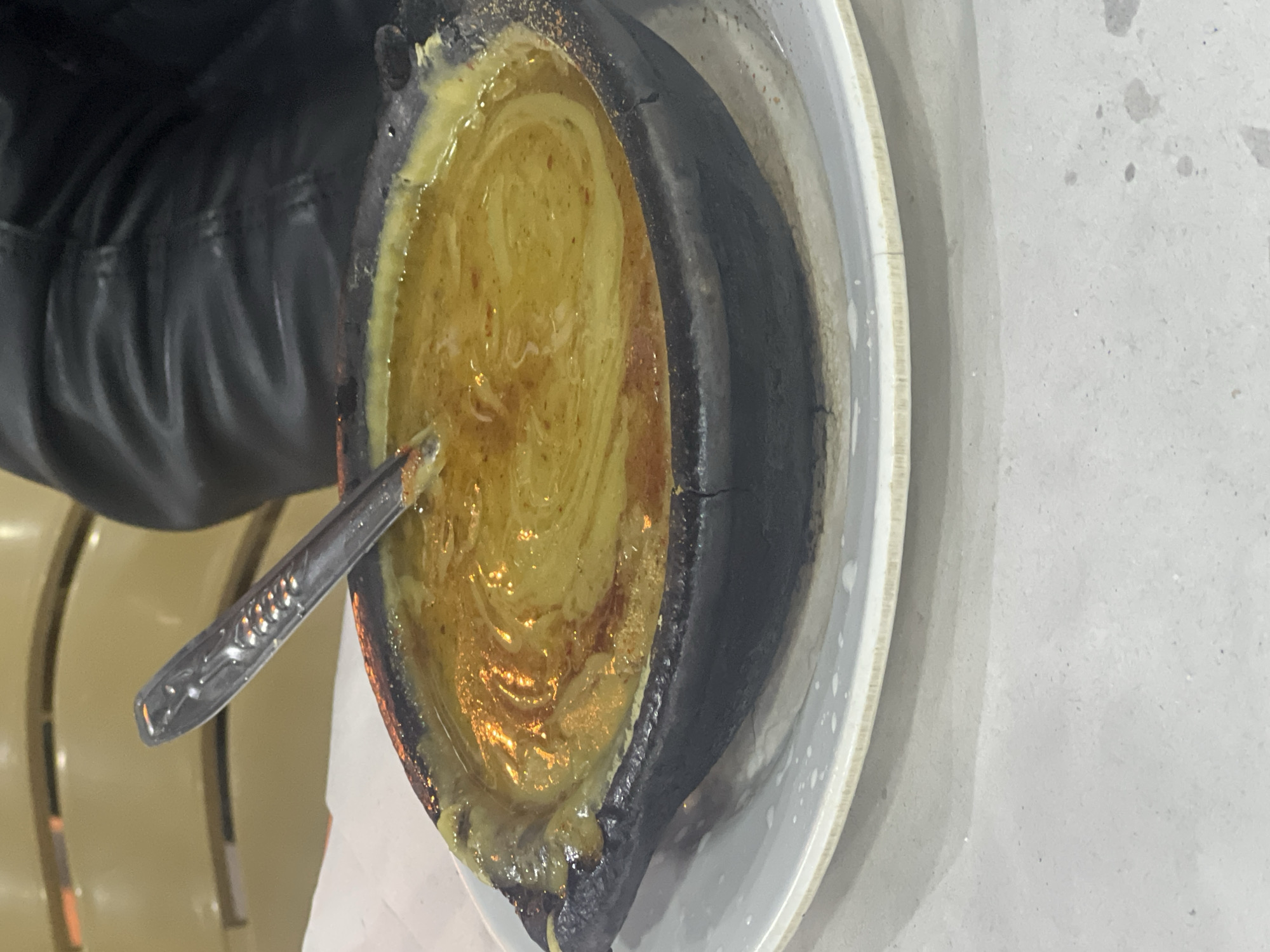 Here is a description of the image:
A close-up, slightly tilted, image shows a dark ceramic tajine dish filled with a yellowish, oily sauce. A silver spoon rests in the dish. The tajine sits on a white plate. To the left, the leg of a person wearing a black leather jacket can be seen. In the background, portions of a light-colored wicker chair are visible, and the surface beneath the plate appears to be a light gray concrete. The photograph was likely taken in M'Diq, Morocco.