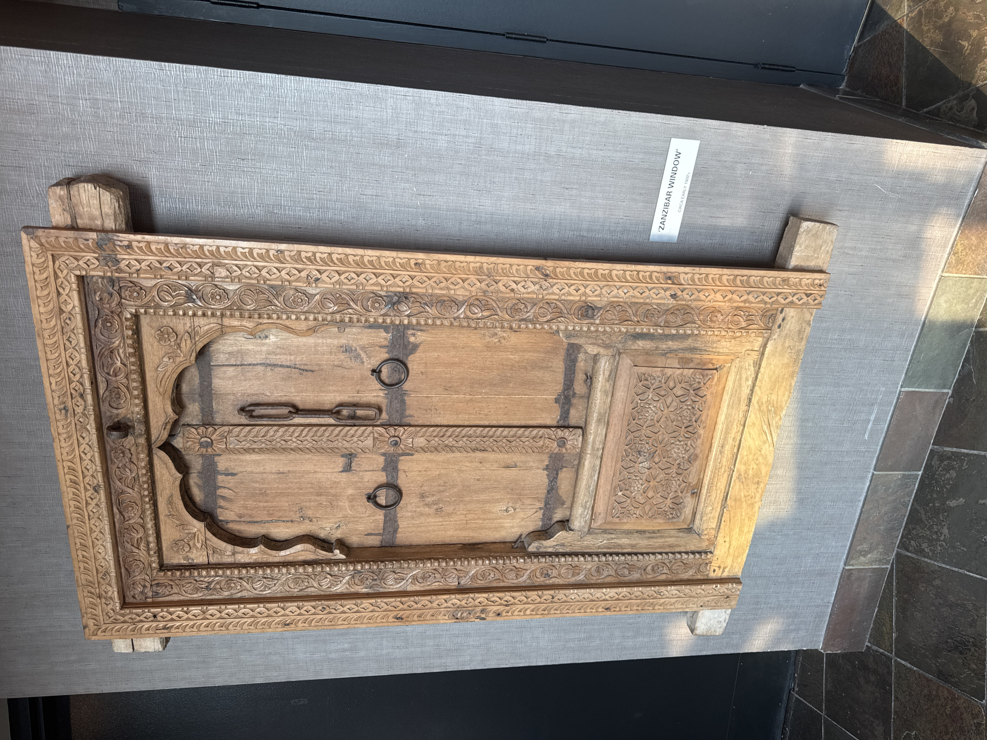 A wooden, antique window frame with intricate carvings and two small wooden doors is mounted on a textured grey wall in a dimly lit room. The frame, measuring approximately 3 feet by 2 feet, displays detailed floral and geometric patterns. The doors are secured by two metal chains and feature round metal handles. Below the doors, a carved panel with geometric designs is visible. The window frame is supported by two rough-hewn wooden beams that extend from the sides of the frame to the wall. A small rectangular plaque is affixed to the wall to the right of the window, reading "'ZANZIBAR WINDOW' CIRCA EARLY 1900'S". The floor is tiled with large, dark, irregular-shaped stones. The environment suggests an indoor exhibition or display space, possibly in the city of North Vancouver, Canada, given the informational plaque.