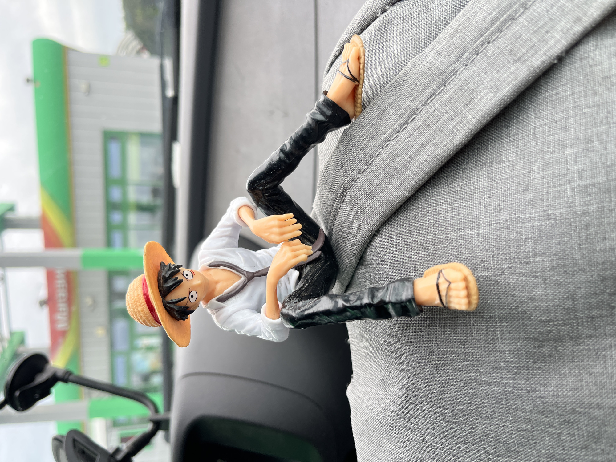 A close-up shot, taken from inside a vehicle, features a Monkey D. Luffy action figure seated on the gray fabric of the car's interior. The figure is depicted with its signature straw hat, white shirt, dark tie, and black pants, with its legs crossed and feet in simple sandals. In the background, out of focus, is a gas station with a distinctive colorful awning and the word "Маяжн" visible on its facade. The sky appears overcast. The location is identified as Shchomyslitski Sel'ski Savet, Belarus.