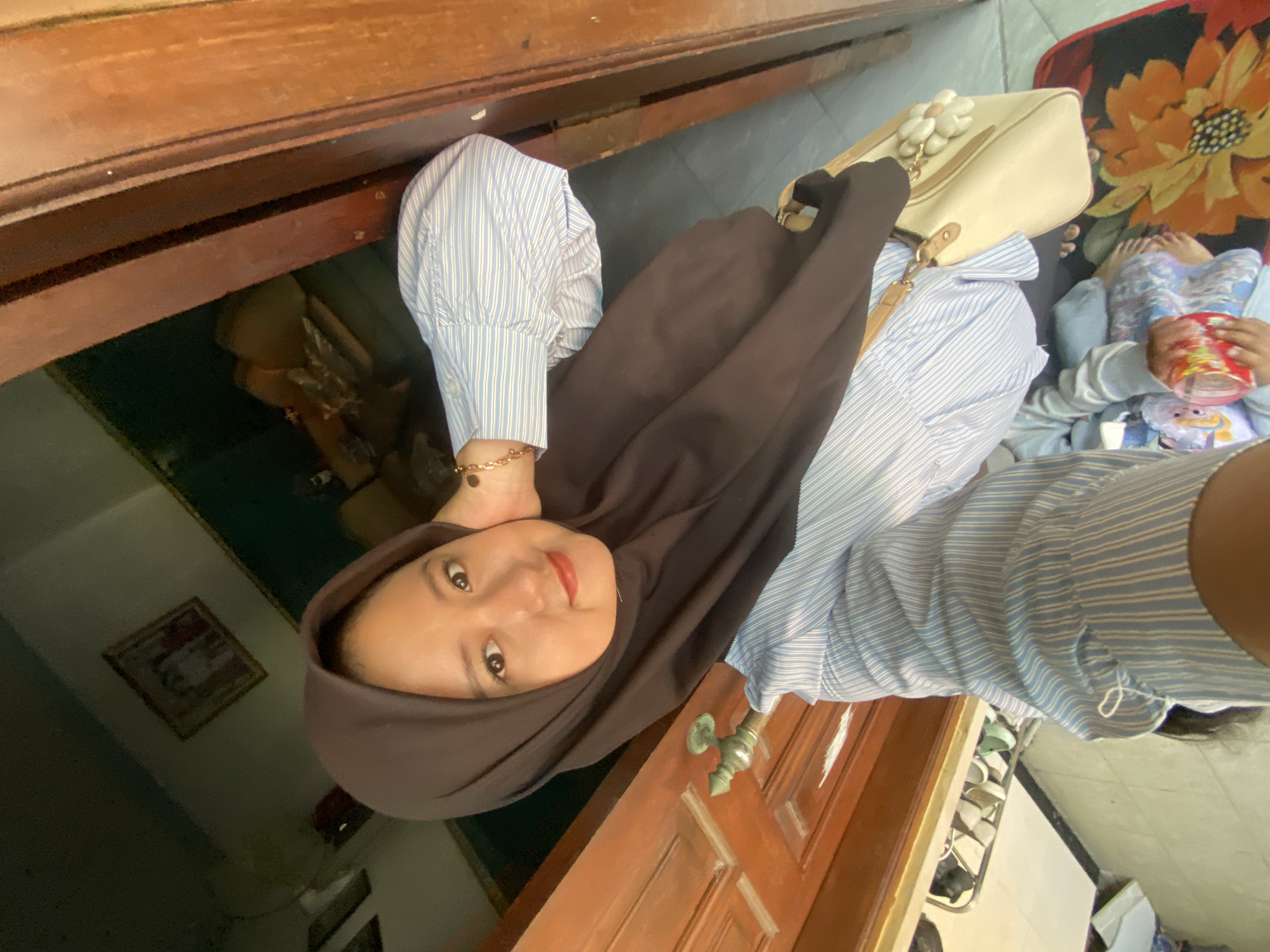 A young woman wearing a dark brown hijab and a blue and white striped long-sleeved shirt poses for a selfie. She is standing in front of a wooden door, with her hand resting on her neck. A light-colored handbag with a floral ornament hangs from her shoulder. In the background, a framed picture is visible on the wall, and part of a shoe rack can be seen. The scene appears to be an interior residential setting in Gelam, Indonesia.
