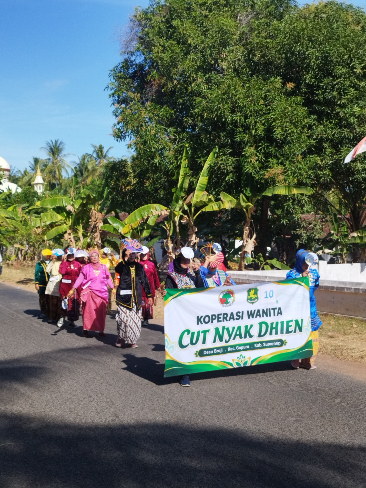 In Andulang, Indonesia, a group of women are participating in a parade. They are walking down a paved road lined with trees and vegetation. Some of the women are wearing traditional Indonesian attire, including sarongs and colorful tops, while others have on headscarves. They are holding fans and a banner that reads "KOPERASI WANITA CUT NYAK DHIEN" and "Desa Braji, Kec. Gapura, Kab. Sumenep." The sky is clear and blue, and the sun is shining, casting shadows on the road. In the background, there are glimpses of buildings, possibly a mosque, and more palm trees, suggesting a tropical or sub-tropical setting. The overall atmosphere appears festive and communal.