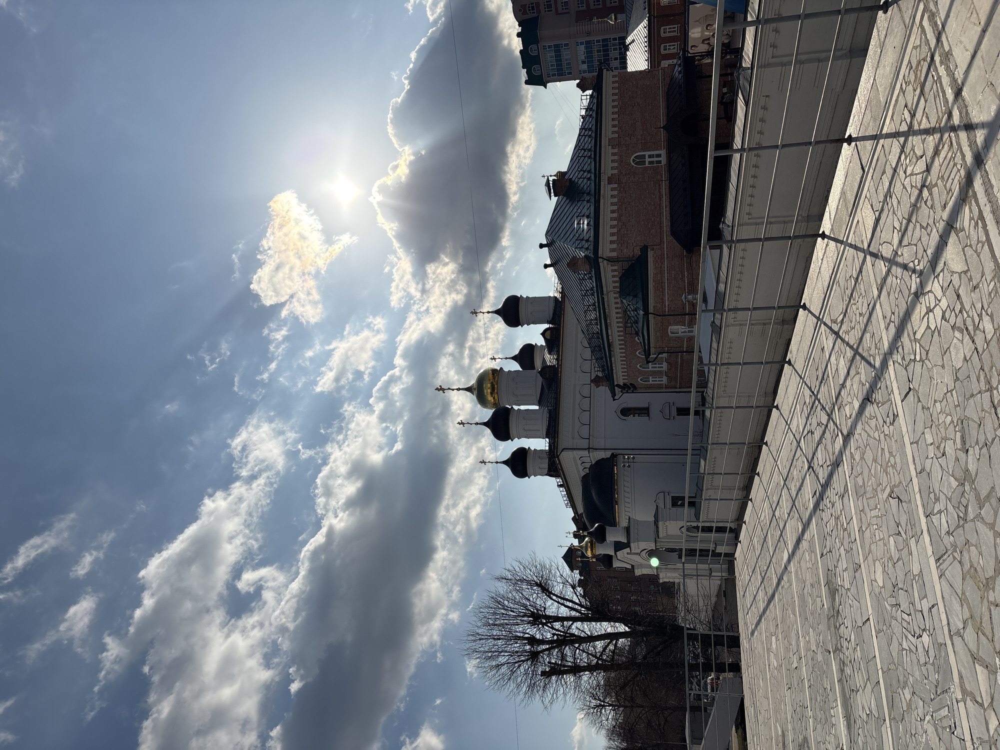 The image depicts a view of the city of Ulyanovsk, Russia, capturing a portion of the cityscape and the sky above. The scene includes a white building with black and gold domes, characteristic of Russian Orthodox architecture. A building with orange brick facade is present behind the Orthodox church. The sky is partially covered with clouds, with the sun shining brightly through some of them. A tree with bare branches is also in the foreground. A structure with light gray, fragmented stone or tiled surface is at the right corner. The image is taken from a low angle, looking up at the buildings and the sky.
