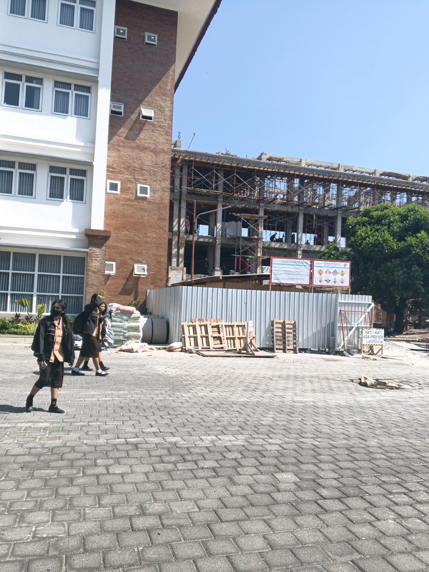 The image shows a street scene with a modern building on the left and a construction site on the right, under a clear blue sky. The building on the left is white with multiple floors, featuring rows of windows with blinds. Adjacent to it is a section of brick facade with smaller square windows. In front of the building, three individuals, possibly students in uniform, are walking on a paved walkway. They are wearing dark jackets over light-colored shirts and dark skirts or shorts. One person in the foreground is wearing a black mask.
The construction site on the right is characterized by scaffolding and exposed concrete structures, indicating ongoing building work. A tall green tree is visible behind the construction site. A corrugated metal fence separates the paved area from the construction zone. Several stacks of wooden pallets and bags of cement are scattered around the site, along with some construction materials and debris. Two informational signs are posted on the fence within the construction area. The ground is paved with gray interlocking bricks. The overall scene suggests an urban environment in Denpasar, Indonesia, possibly near an educational institution given the attire of the pedestrians and the building's architecture.