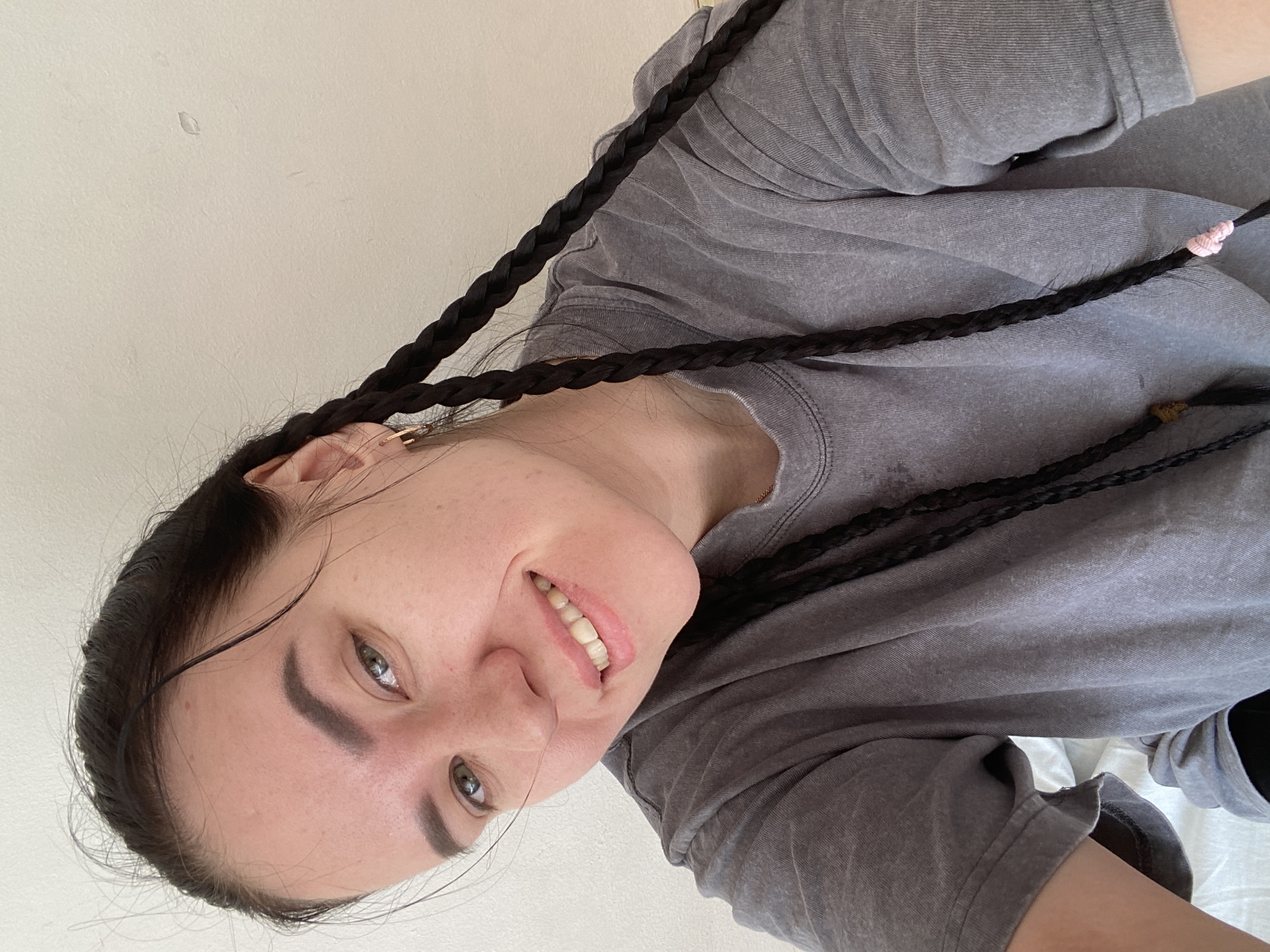 A woman with dark hair braided in pigtails, one secured with a pink hair tie and another with a brown hair tie, is facing the camera. She is wearing a grey crew neck shirt and is smiling. The background is a plain white wall. The scene appears to be indoors. There is no information to confirm the location as Perm, Russia, therefore I cannot include that information.