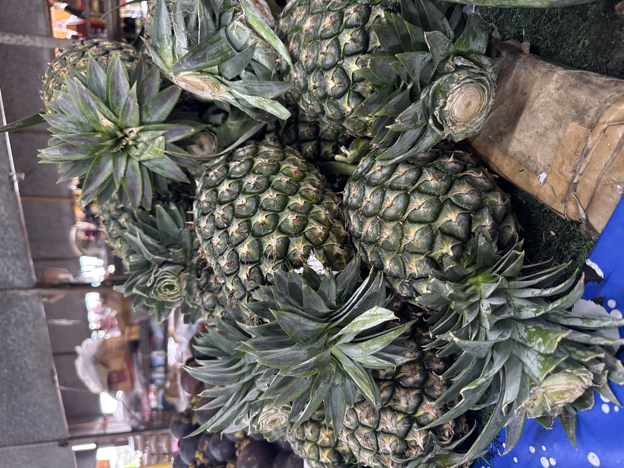 A close-up, eye-level shot focuses on a pile of fresh pineapples with their green crowns. The pineapples have rough, segmented skins and are arranged in a jumbled heap. In the background, out of focus, are other fruits, possibly mangosteens, and market stalls with various goods. The scene appears to be an outdoor market in Baan Bo Phut, Thailand, with a rough, possibly corrugated metal roof overhead. The lighting is bright, suggesting daytime.