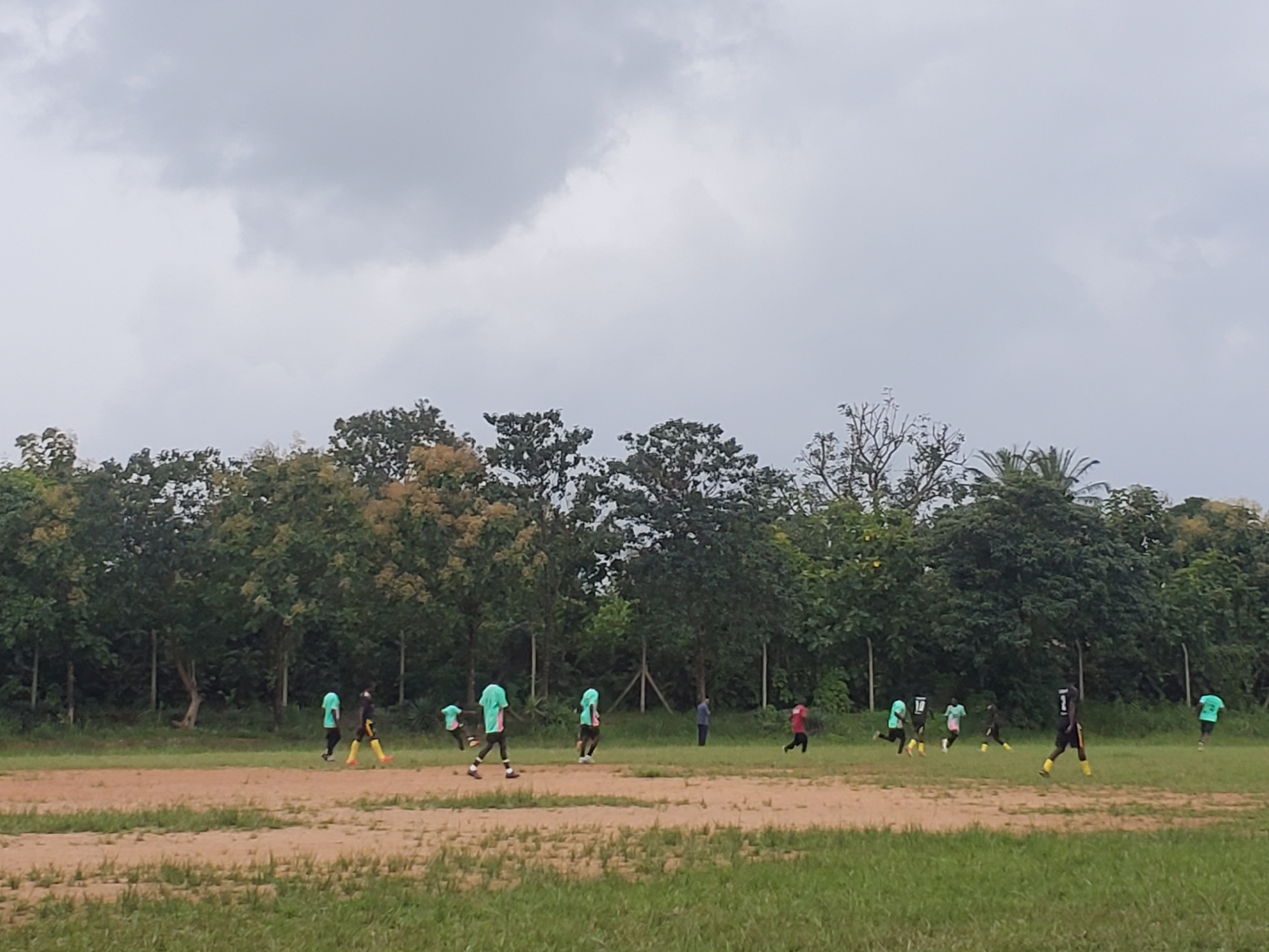 A group of young people are playing soccer on a dirt field in Miango, Nigeria. The sky is overcast and the trees are lush green. The players are wearing various colored shirts and shorts. Some of the players are running, while others are standing. The field is uneven and there are patches of grass scattered throughout.