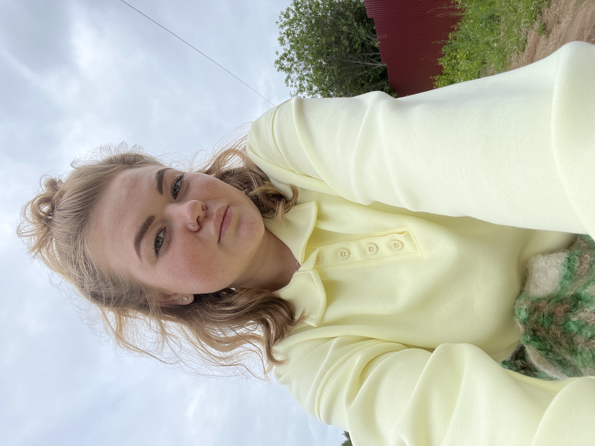 The image depicts a close-up selfie of a young adult female located outdoors in Chaikovsky, Russia, under an overcast sky. The subject, positioned centrally, has light-colored hair styled in a messy high bun with loose, wavy strands around her face. She looks directly at the camera with a neutral expression. Her left ear displays three small stud piercings, and a silver-toned hoop piercing is visible in her left nostril. She is wearing a pale yellow, long-sleeved, collared sweatshirt with a four-button placket. A glimpse of a multi-colored (green, brown, white) patterned fabric, possibly a blanket or another garment, is visible in the lower left portion of the frame. The background consists predominantly of a uniform gray, cloudy sky. In the mid-right, a red corrugated metal fence is present, with a green-leafed tree and sparse vegetation visible behind it. A thin, dark horizontal line, possibly a utility wire, stretches across the upper right quadrant of the sky. The immediate foreground on the right shows a dirt path with some patches of green grass.
