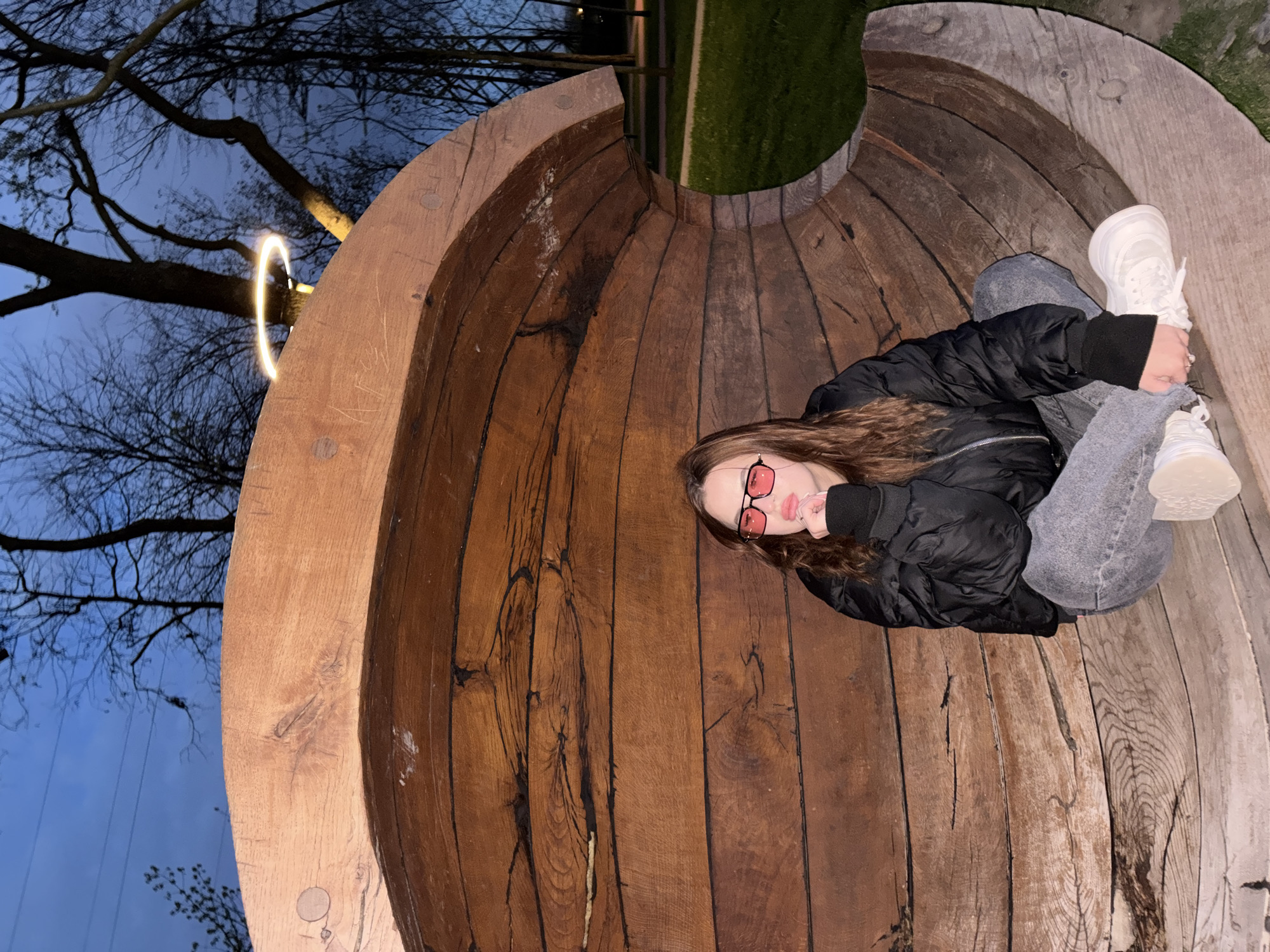 An evening outdoor shot shows a young woman relaxing inside a large, concave wooden structure, likely a public art installation or bench, in Krasnodar, Russia. The structure is composed of numerous vertical wooden planks, forming a bowl or barrel-like interior where she is comfortably situated.
The woman, with long brown hair, is wearing a black puffer jacket, gray jeans, and white sneakers. She sports distinct red-tinted sunglasses and looks directly at the camera with a slight pout, conveying a relaxed and perhaps stylish demeanor.
The scene is set at dusk or night, under a deep blue sky. Bare tree branches are visible against the sky, suggesting autumn or winter. A striking feature is a bright, glowing circular light fixture, resembling a neon ring, suspended near one of the trees. In the background, beyond the wooden structure, patches of green grass are discernible. The wooden planks display visible grain, knots, and some signs of wear and tear. There is no clearly discernible text visible in the image.