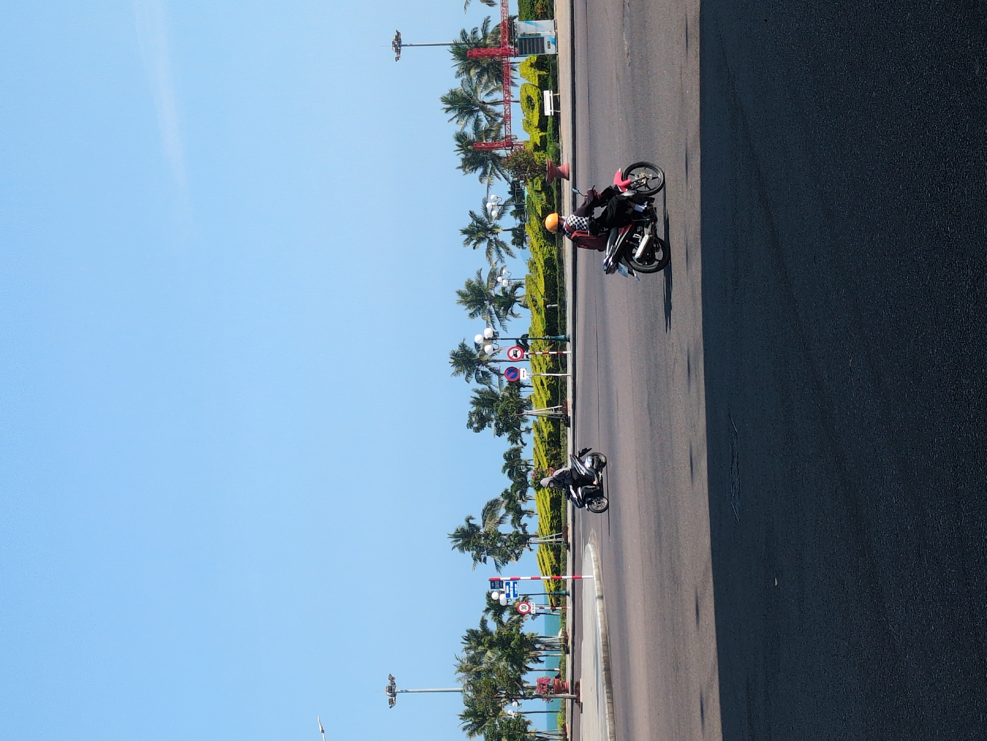 The image depicts a street scene in South Quy Nhon Ward, Vietnam. Two motorcycles are traveling on the road. The road is bordered by a sidewalk with palm trees and manicured hedges. Street lamps and various traffic signs are visible along the edge of the road. The sky is clear and blue.