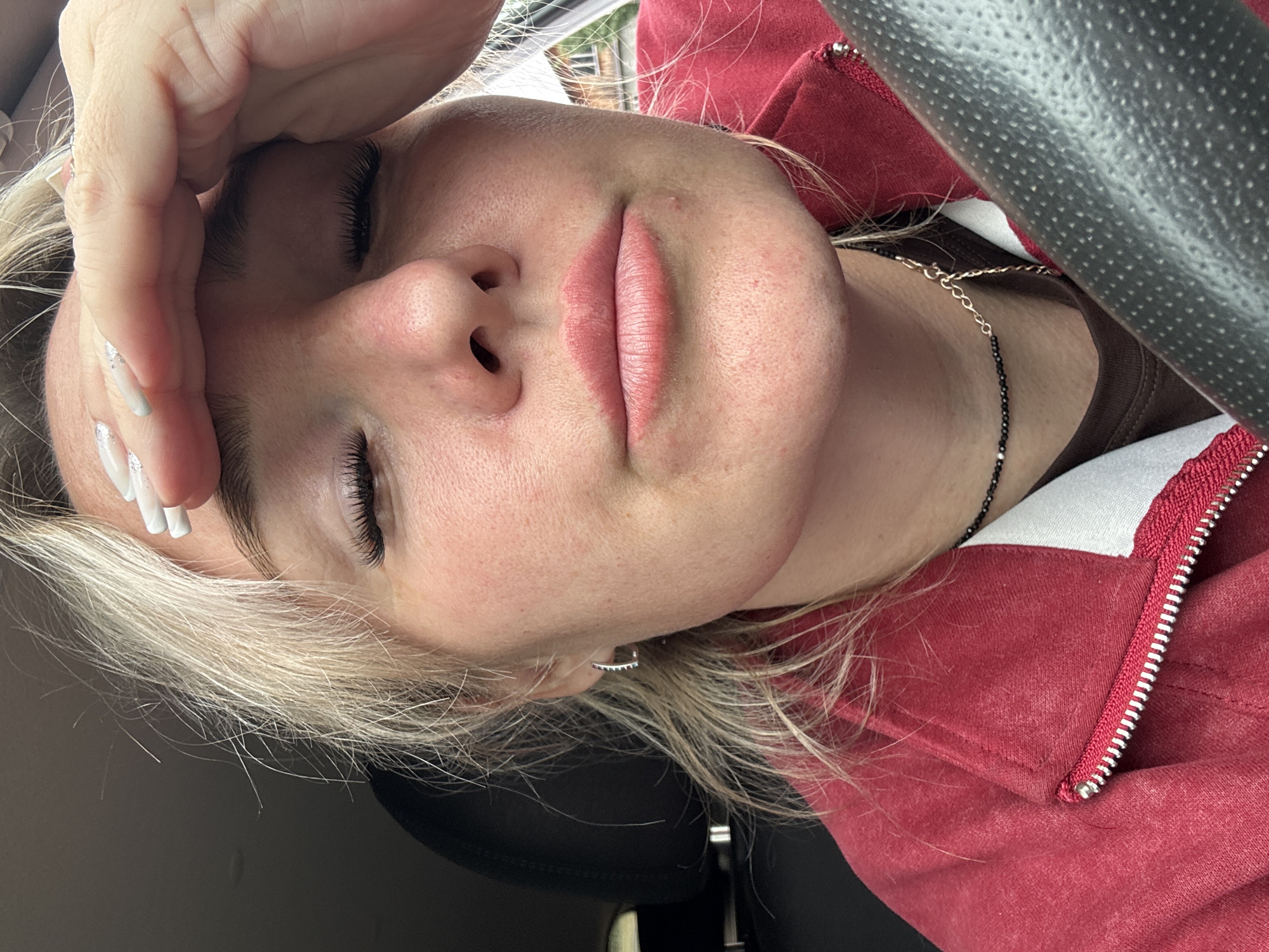 The image is a close-up, waist-up, selfie-style photograph of a young woman inside a vehicle. The woman has fair skin, blonde hair, and is wearing makeup, including false eyelashes. She is wearing a red zippered jacket over a brown shirt, and a necklace with black and gold beads. Her left hand is pressed against her forehead. The interior of the car is visible, including a steering wheel, car seat, and a part of the dashboard. The background is blurred and appears to be outdoors, possibly in a residential area with buildings and trees. The exact location in Chastyye, Russia, cannot be determined from the visual information provided.