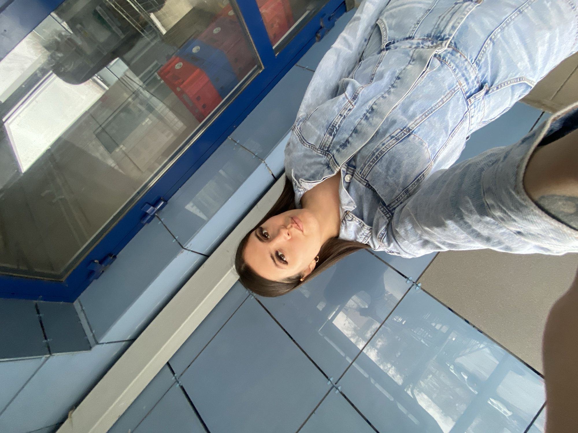 A young woman with dark hair is posing for a selfie in an urban environment. She is wearing a light blue denim jacket and stands next to a blue structure with a window reflecting interior details, possibly crates or containers. The floor is tiled with blue panels, and the overall scene appears to be outdoors, given the daylight and reflective surfaces. The image provides no information about the specific location in Chaikovsky, Russia.