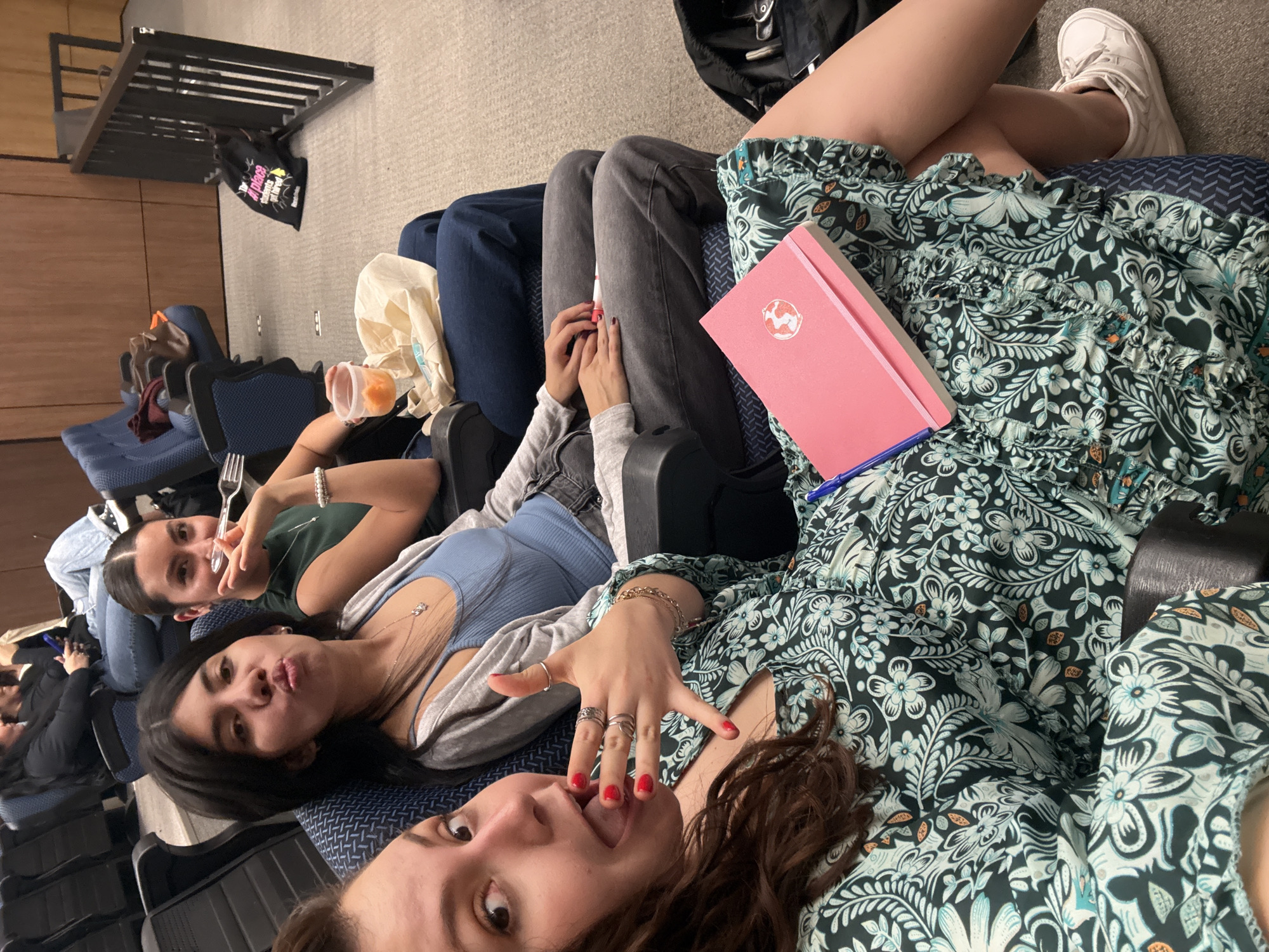 Three young women are lying down in a row of theater seats. The woman closest to the camera has brown hair and is sticking her tongue out. She is wearing a dress with a dark floral pattern and has multiple rings on her hand, with red nail polish. The woman in the middle is puckering her lips and is wearing a blue top with a gray cardigan. The woman in the back is smiling and holding a fork, wearing a green shirt and a bracelet.
A pink notebook with a logo is on the lap of the woman in the floral dress. A white tote bag and a container of fruit are visible near the woman in the green shirt. A black backpack and a pair of white sneakers are visible near the woman in the floral dress.
The theater seats are blue and have a patterned design. A metal rack is visible in the background, along with a black tote bag. The location is identified as Mexico City, Mexico.