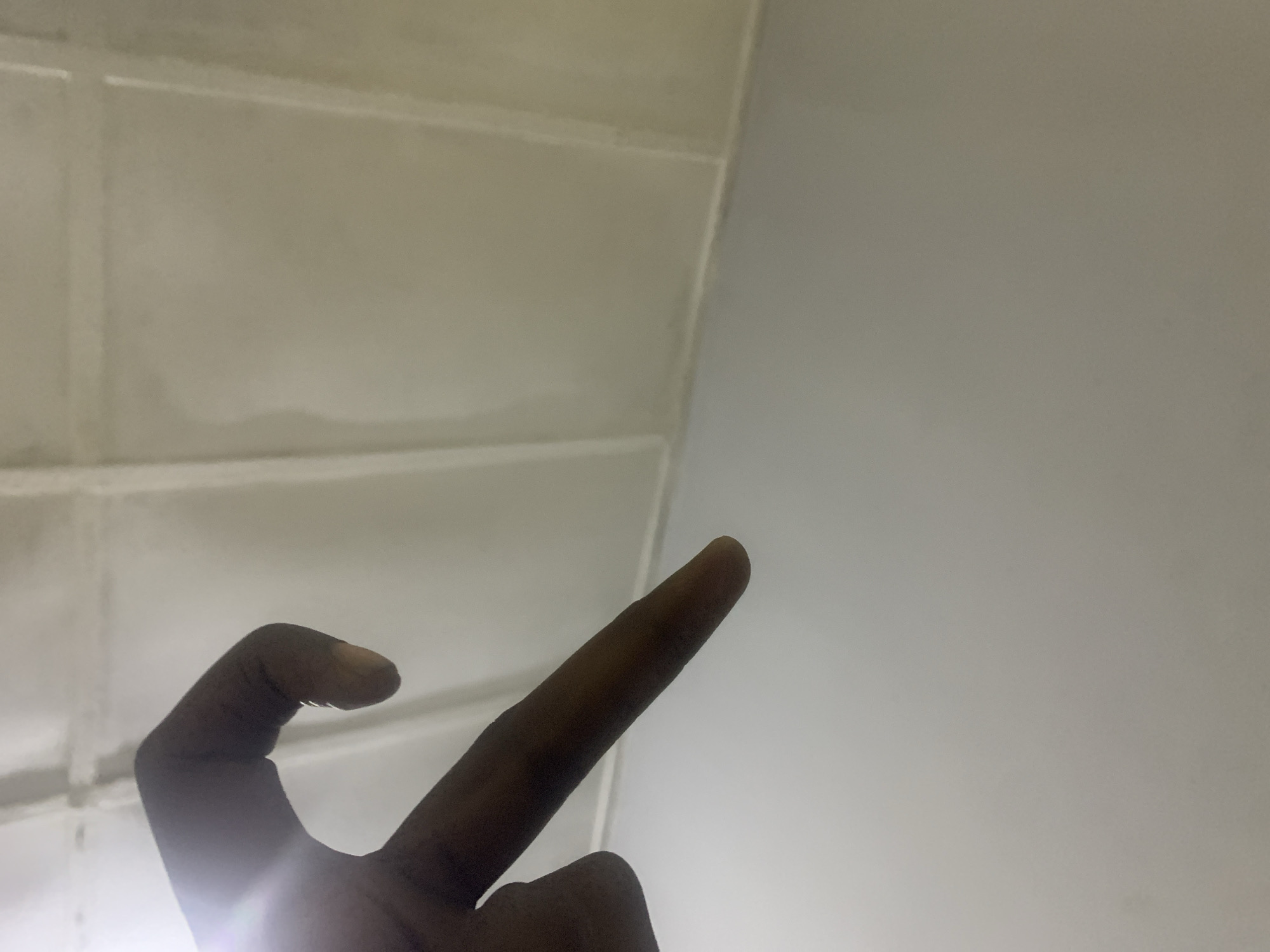 The image shows a dark-skinned hand with the index finger extended and other fingers bent. The hand is positioned in front of a background consisting of white, rectangular tiles on the left and a plain, light-colored wall on the right. The lighting appears to be artificial and somewhat diffused, casting a slight glare on the hand. No specific details indicate the location is Rafin Par, Nigeria.