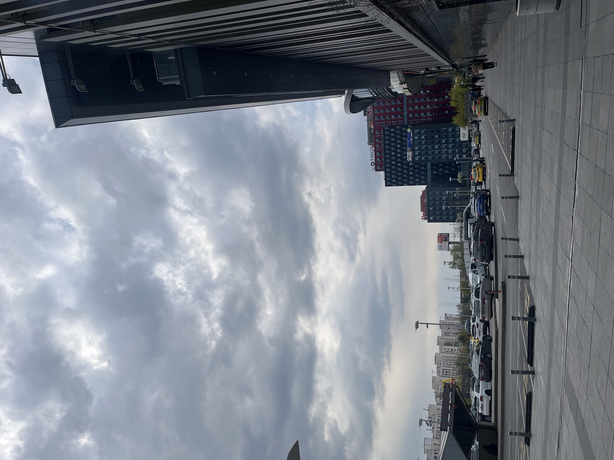 The image, correctly oriented, displays a modern urban street scene in Sofia, Bulgaria, under an overcast sky. The viewpoint is from beneath a large, dark, contemporary building's overhang, which occupies the upper left section of the frame, showcasing its sleek design and integrated lighting.
Across a wide, tiled pedestrian area and a street, a busy urban environment unfolds. A row of parked vehicles, including several bright yellow taxis, lines the right side of the road, with additional cars visible further down the street. In the mid-ground, several multi-story buildings rise against the dense cloud cover. One distinctive building features a facade of red and dark sections with numerous windows, bearing the text "TINGIN OZON" and "SGS-RENU". Another dark building is partially visible behind it, with more city structures receding into the background.
A few indistinct figures of people can be seen walking on the sidewalk adjacent to the parked cars. The sky is dominated by thick, grey clouds, indicating an overcast or cloudy daytime, though sufficient ambient light is present. A "DO NOT ENTER" sign is partially visible near the street level in the distance. The overall scene captures typical daily activity within a modern city setting.