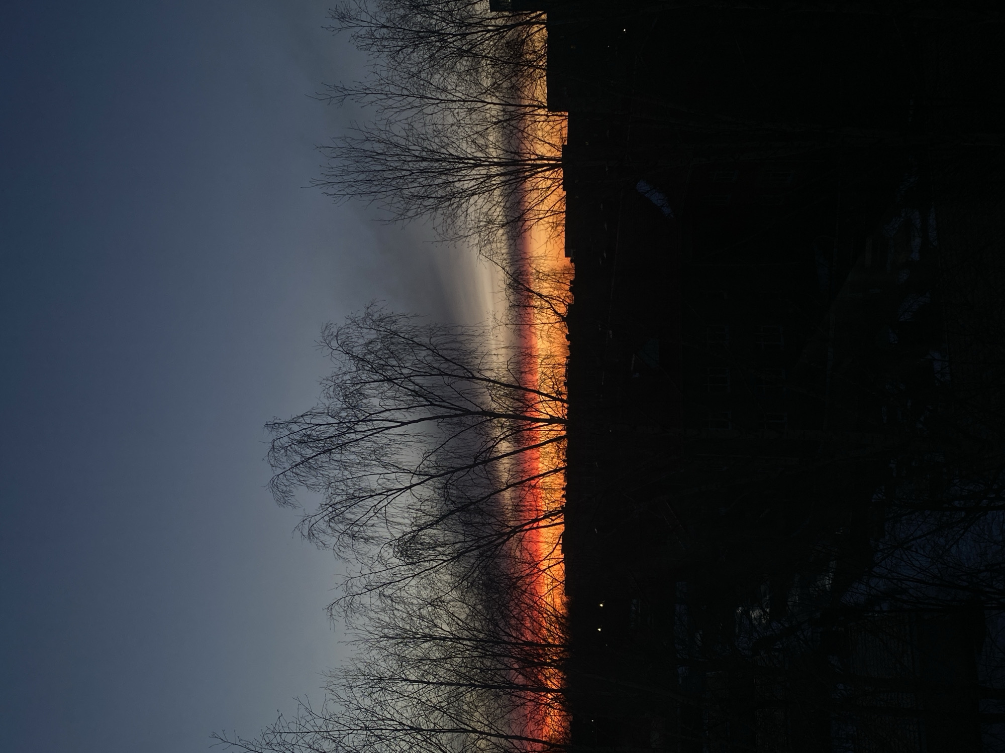 The image shows a sunset scene in Izhevsk, Russia. The sky transitions from a dark blue-gray in the upper left to orange and red hues near the horizon. Silhouetted bare trees are visible in the foreground, with some branches reaching into the sky. In the background, dark shapes suggest buildings and structures characteristic of an urban environment. Two small, indistinct light sources may be streetlights or windows of buildings. Snow is lightly present on the ground.
