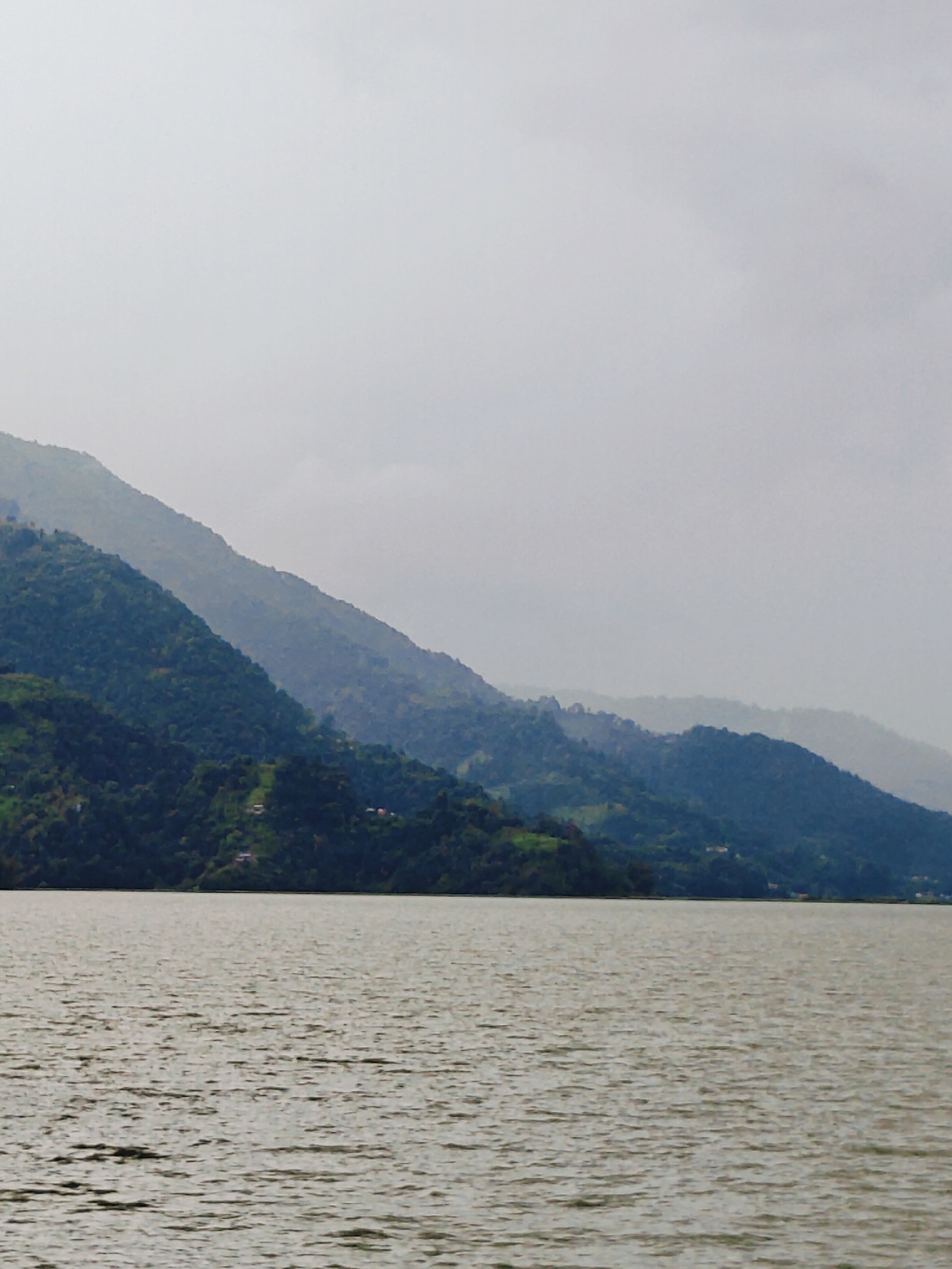 A panoramic view of the Seti River in Pokhara, Nepal, under a hazy, overcast sky. The river dominates the foreground, its surface rippled by a gentle breeze, reflecting the muted tones of the sky. In the mid-ground, lush green hills rise steeply from the water's edge, their slopes dotted with sparse clusters of buildings, suggesting a rural or semi-urban settlement. The hills are layered, with successive ranges fading into the misty distance, creating a sense of depth and vastness. The overall atmosphere is serene and tranquil, with a touch of melancholic beauty due to the overcast weather.