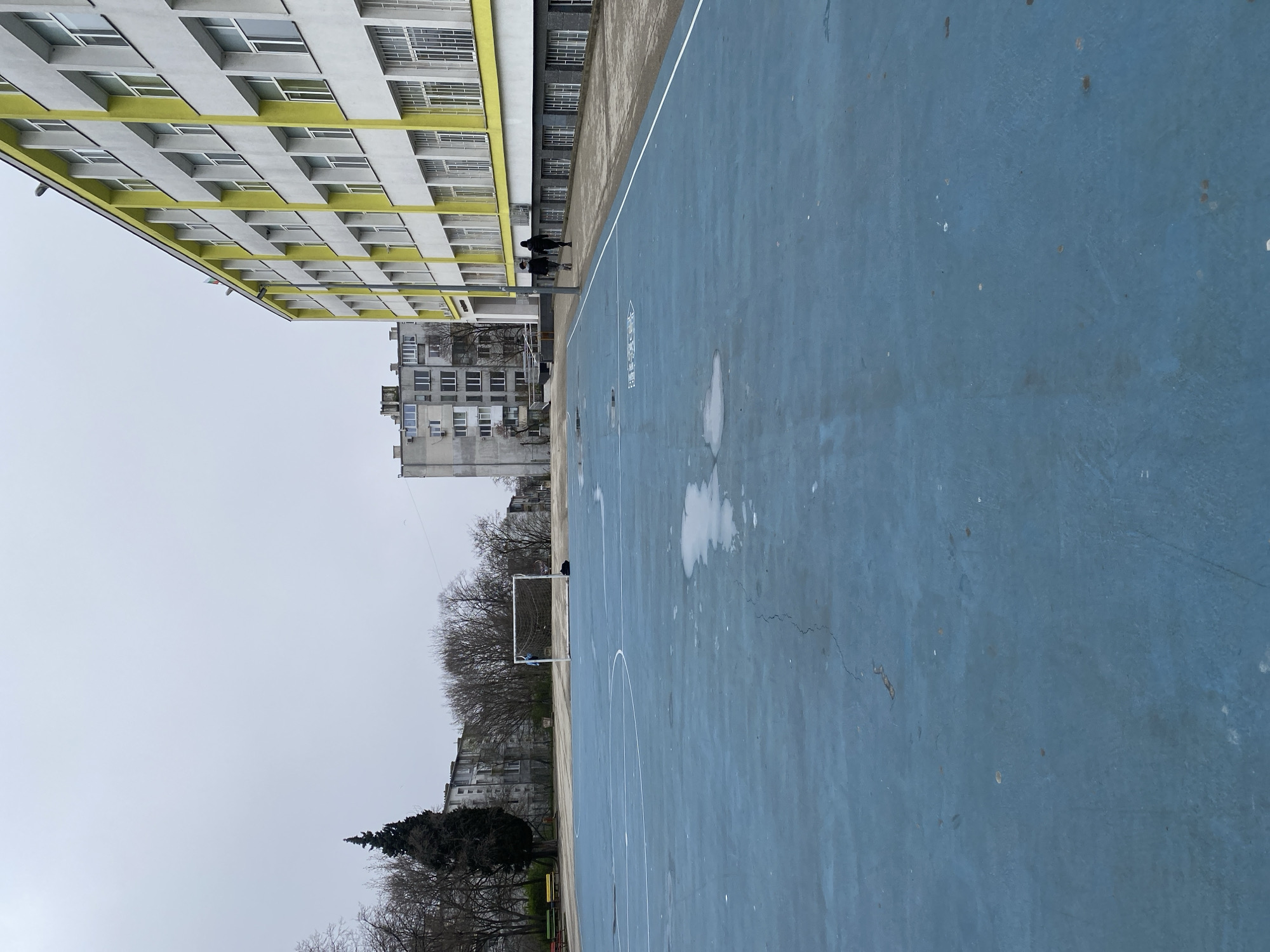 The image depicts a blue outdoor basketball or multi-purpose sports court in Varna, Bulgaria. The court has white lines marking its boundaries and possible game areas. There are patches of moisture on the court surface. A metal soccer goal is visible beyond one end of the court. Several buildings are in the background, including a long yellow and white apartment building. The sky is overcast. Two figures are walking along the side of the court near the building. Bare trees are also visible.