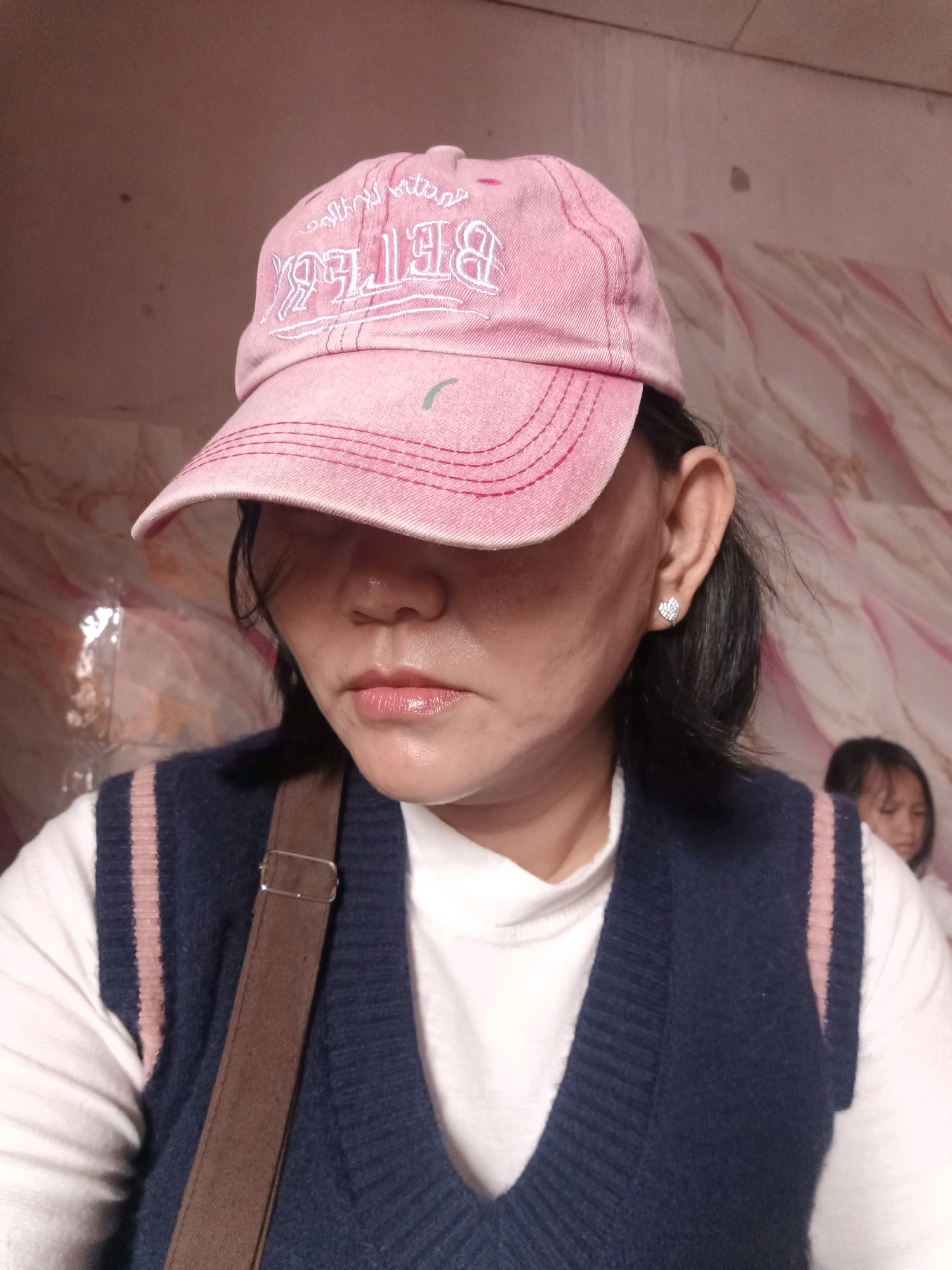A medium shot captures a woman in her 30s or 40s wearing a pink baseball cap, a dark blue vest over a white long-sleeved shirt, and a brown strap diagonally across her chest. The cap's brim partially obscures her face, but her eyes are downcast. She has dark hair, and a small, heart-shaped earring is visible in her left ear. The background consists of a wall with a patterned texture, appearing to be in an indoor setting. In the background, a young girl can be seen slightly out of focus. The image appears to be taken in a residential setting in South Jakarta, Indonesia, based on typical interior design elements.
