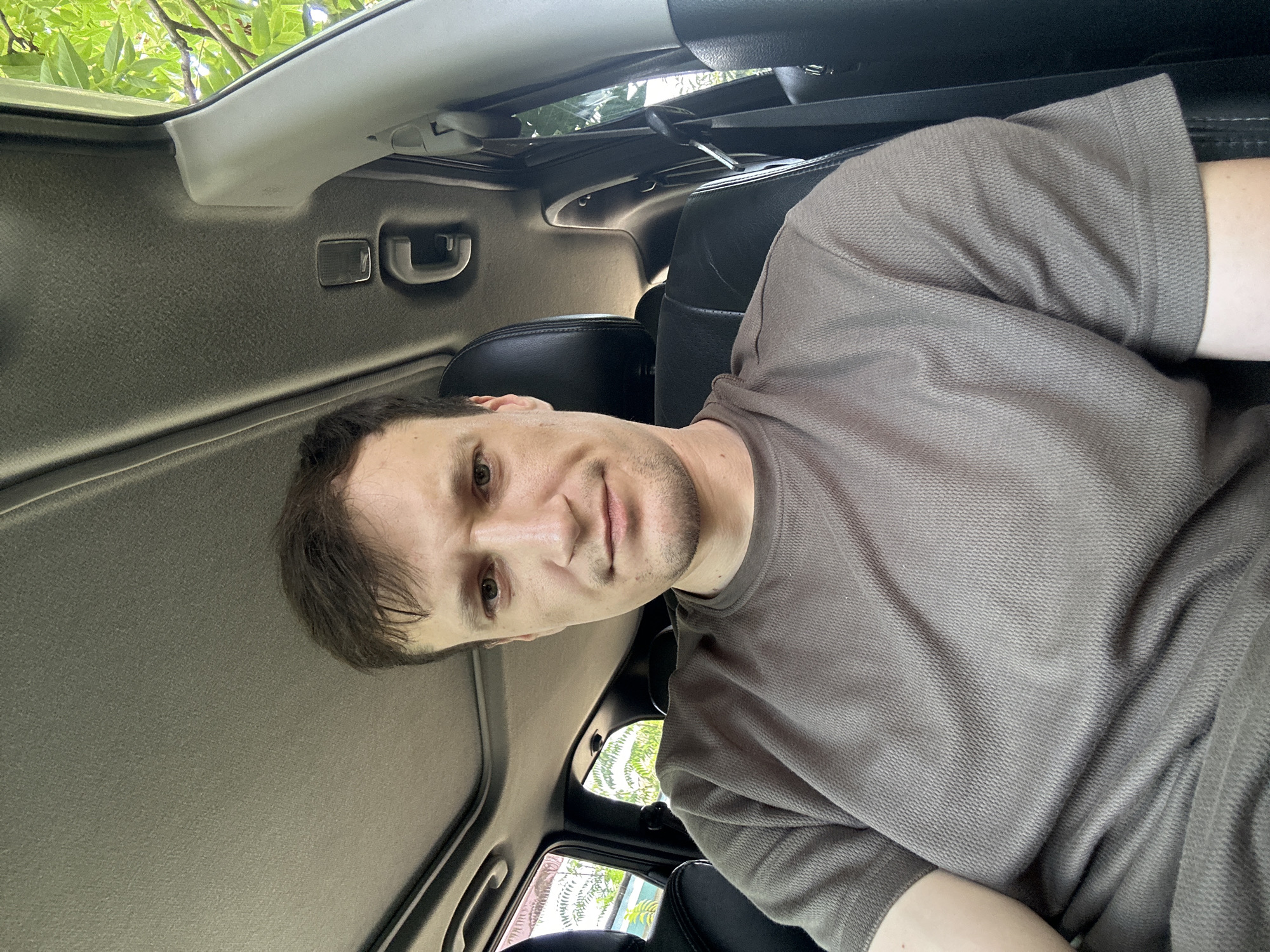 A medium shot captures a young adult male with short, brown hair and a light beard, seated in the driver's seat of a vehicle. He is looking directly at the camera with a neutral expression. The man is wearing a dark gray, short-sleeved shirt with a textured fabric. The interior of the car is visible, including the dark-colored headrest of the seat behind him, the car's ceiling with a sunroof, and a grab handle. Green foliage from trees is visible through the open windows and sunroof, suggesting an outdoor setting, likely in the city of Makiv, Ukraine. The lighting is natural and appears to be daylight.