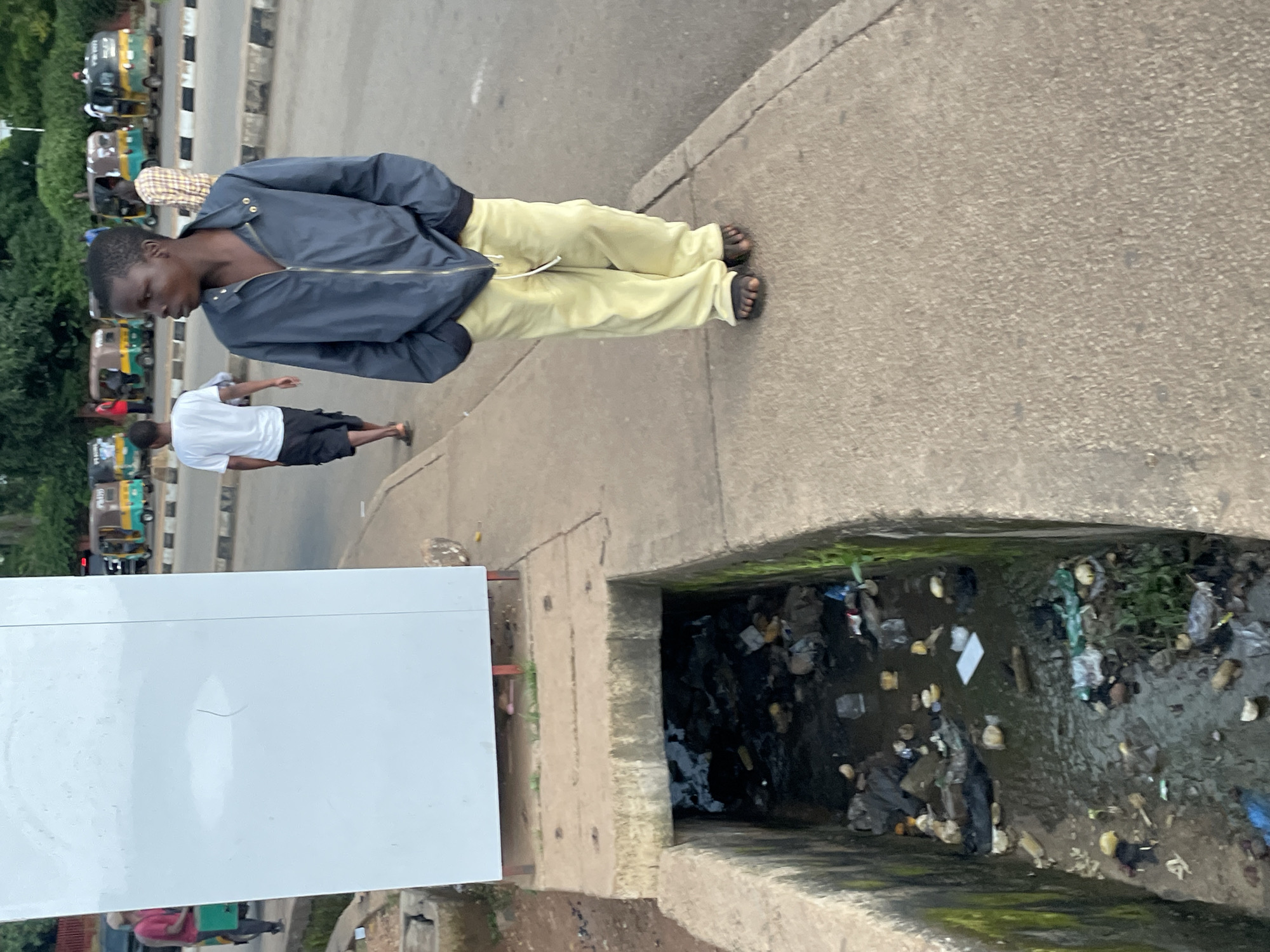 A young Black male stands on a concrete sidewalk, his hands in the pockets of a dark blue zippered jacket and pale yellow pants. He is barefoot, wearing dark flip-flops. His gaze is directed to the right, away from the camera. To his left, a concrete drainage channel is filled with dark water and debris, including plastic bags, bottles, and other refuse. Behind the boy, a white rectangular structure stands on stilts, its purpose unclear. In the background, a street is visible with several three-wheeled vehicles, known locally as Keke Napep, and other pedestrians. The scene suggests an urban environment, possibly in Jos, Nigeria, characterized by its infrastructure and the presence of everyday life.