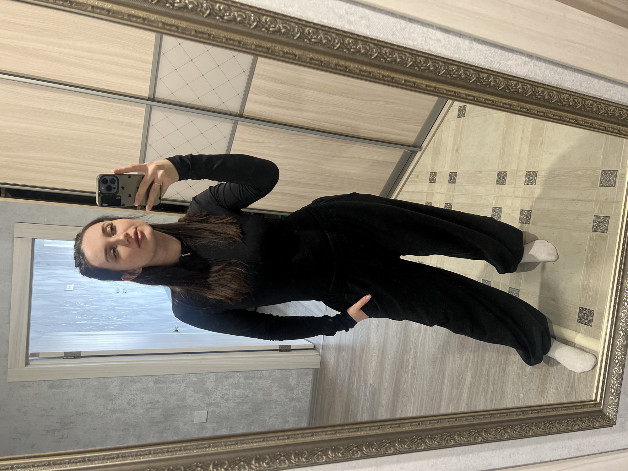 A person, possibly female, is reflected in a mirror. They are wearing a black long-sleeved shirt and loose-fitting black pants, along with white socks. The person is holding a smartphone and appears to be taking a selfie.
The background includes:
- A closet with light-colored panels.
- Flooring of two types: light gray wood-look planks and a patterned light-colored tile.
- A door and wall are visible.
There is not enough information to determine that the location is in the city of Tyumen, Russia.