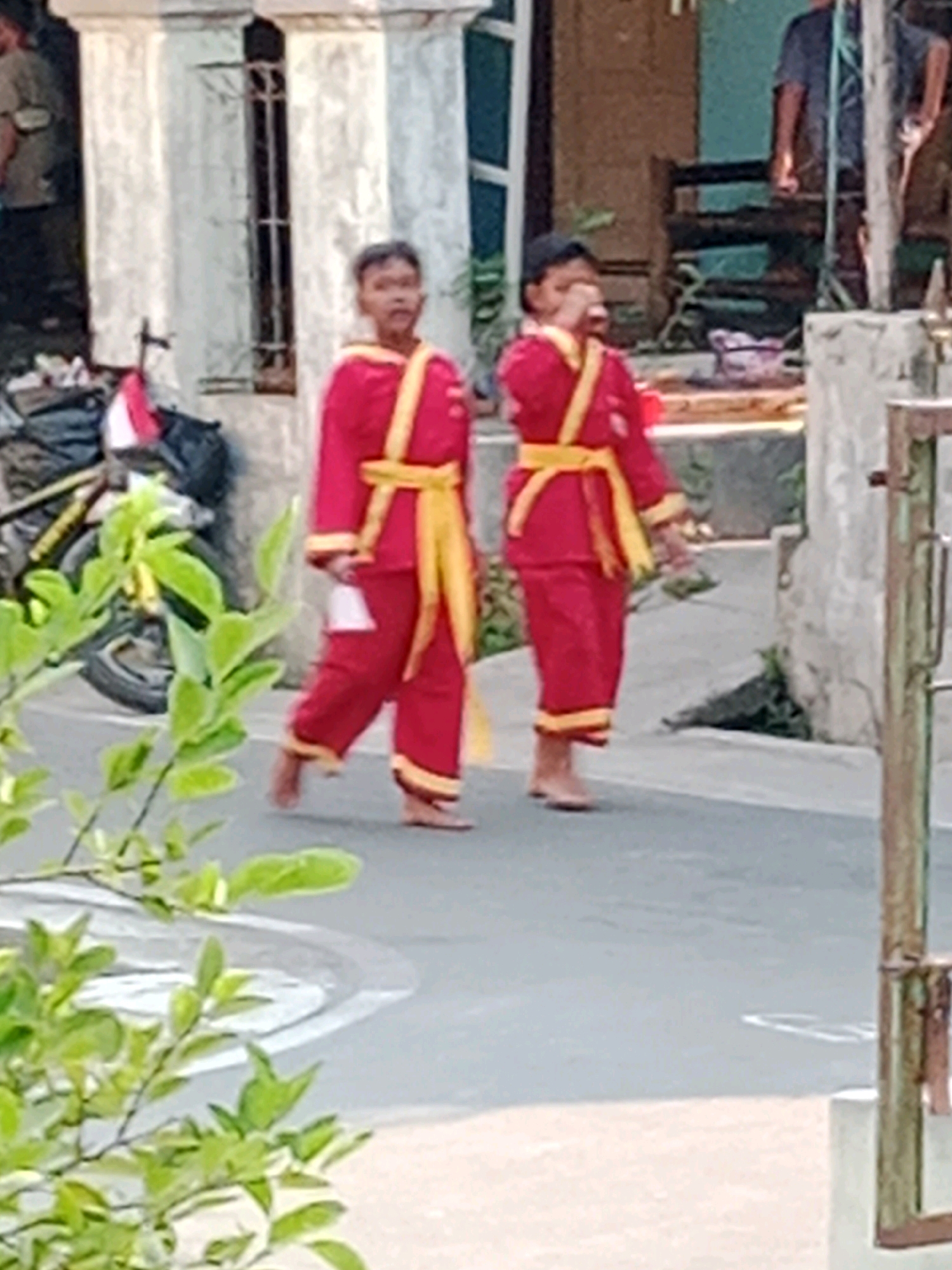 The image shows two young boys dressed in red martial arts uniforms with yellow sashes, walking on a paved street in Waru, Indonesia. The boy on the left is looking towards the camera, while the boy on the right is looking down. They appear to be participating in a parade or event, as indicated by their matching attire. In the background, there are buildings with white pillars and a bicycle. The overall scene is outdoors, with greenery visible in the foreground.