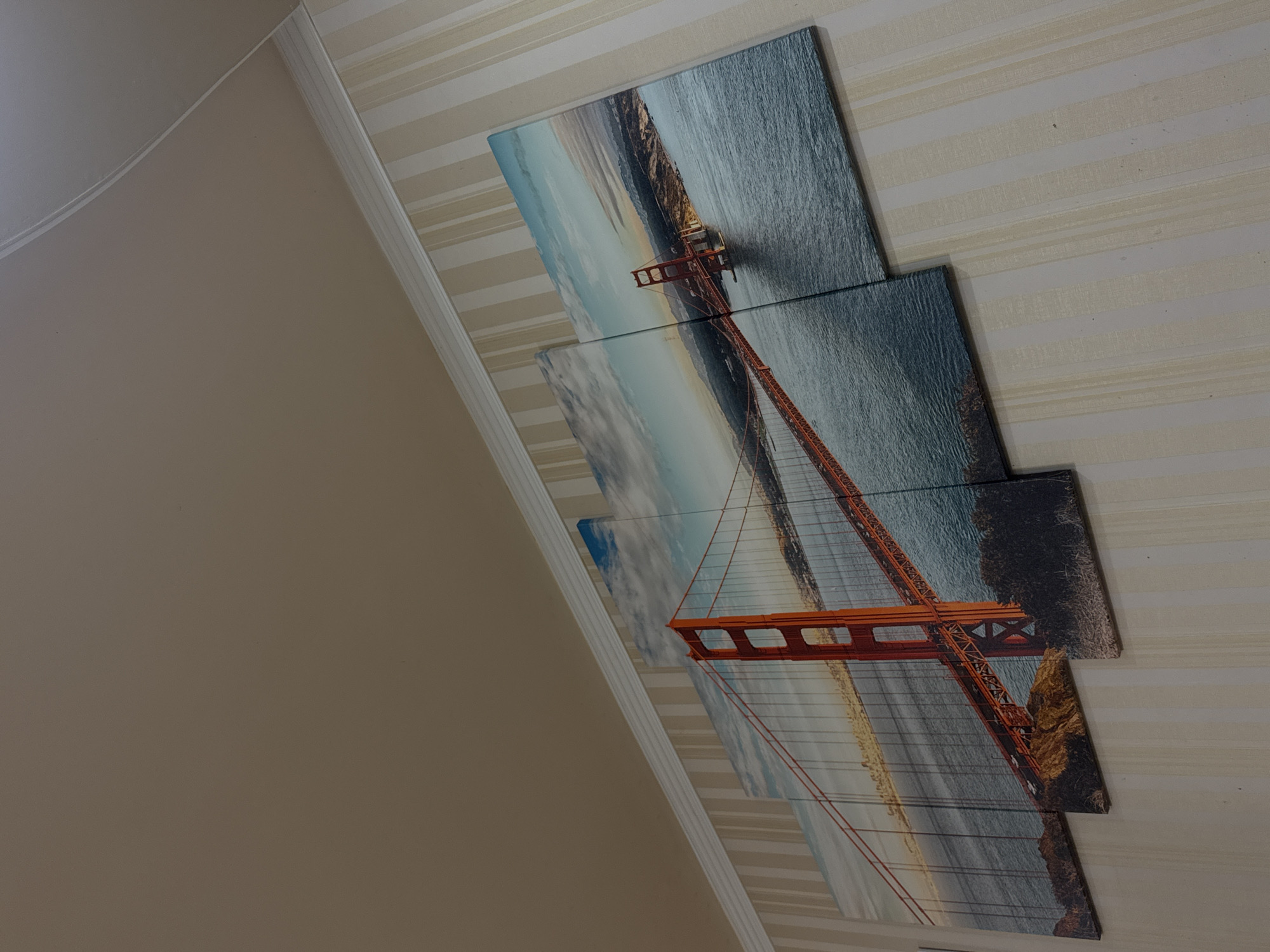 A close-up shot taken from an off-kilter angle depicts three wall-mounted artwork pieces, each featuring sections of a panoramic view of what appears to be the Golden Gate Bridge. The pieces align to create a continuous image of the bridge spanning across a body of water. The artwork is displayed against a striped wallpaper background. The location and information about the city of Yerevan, Armenia are not discernable from the provided image.