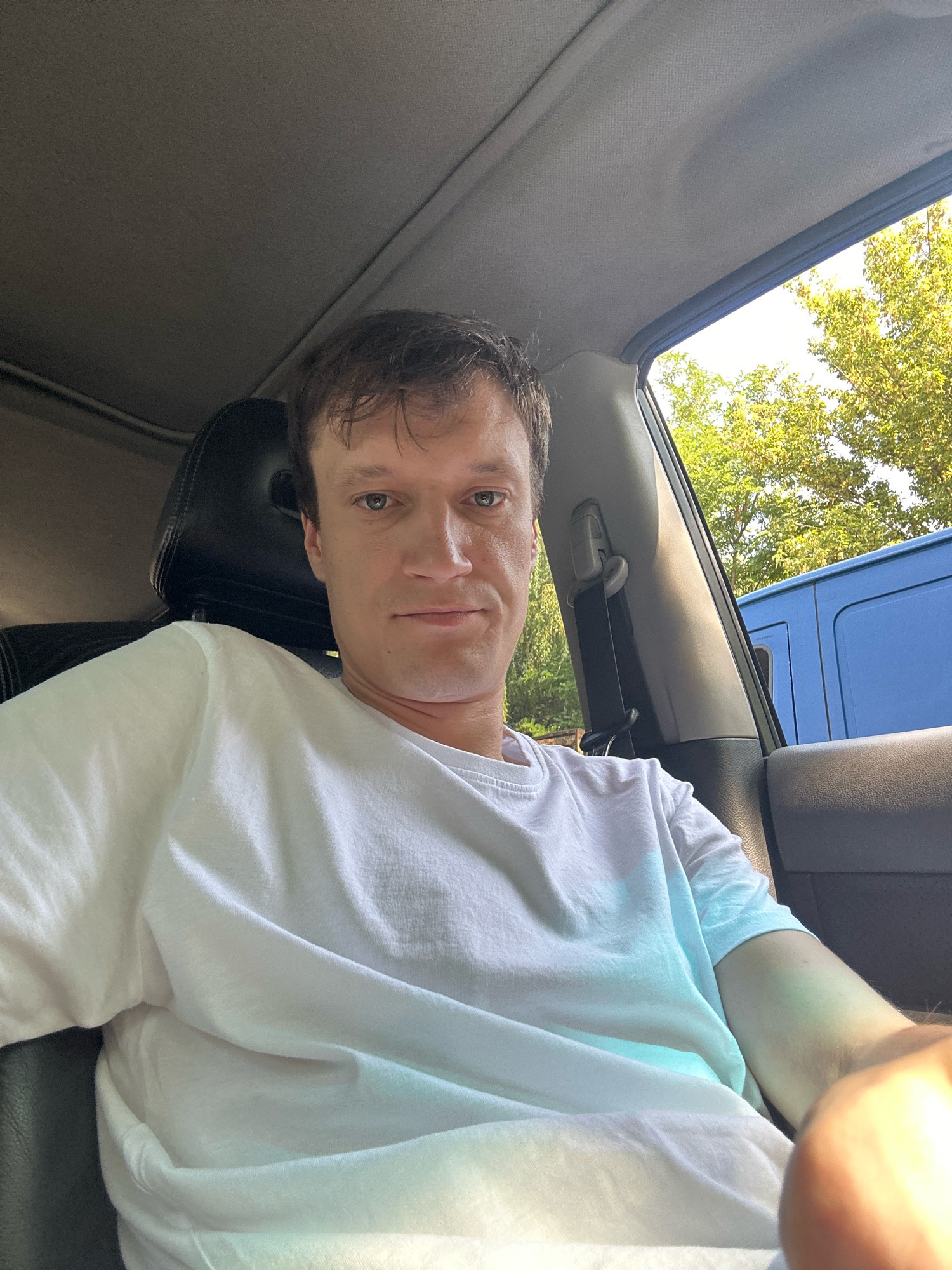 A man with short brown hair and blue eyes is sitting in the driver's seat of a vehicle, taking a selfie. He is wearing a white t-shirt and looking directly at the camera. The interior of the car is visible, including the headrest of the passenger seat and a seatbelt. Outside the car, green trees and a blue van are visible under a bright sky, suggesting an outdoor setting in Kamianets-Podilskyi, Ukraine.