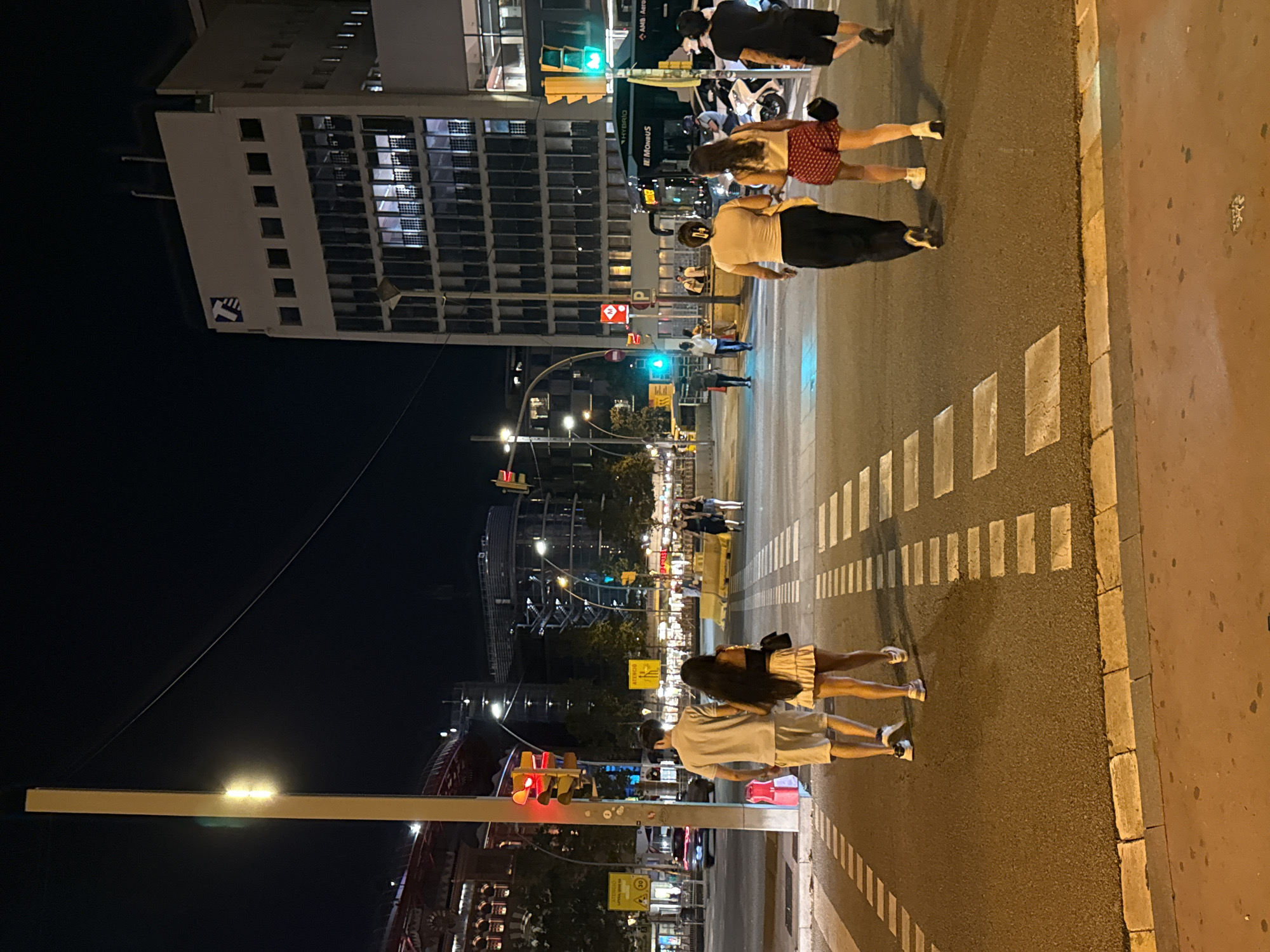 The image shows a nighttime street scene in Barcelona, Spain. In the foreground, a couple walks across a crosswalk, holding hands. The man wears a light-colored polo shirt and shorts, while the woman wears a black top and a ruffled skirt. Behind them, other pedestrians are visible, as well as traffic lights and streetlights illuminating the asphalt.
In the mid-ground, a green bus with the text "MONBUS" and "HYBRID" is stopped at a bus stop, alongside motorcycles and scooters. Further back, a modern building with numerous windows and balconies stands tall. The architecture suggests it could be an office building or residential complex. A prominent red "M" sign with a circle, likely indicating a metro station, is also visible.
The background features more urban structures, including what appears to be a stadium or large entertainment venue with red accents, and scaffolding on another building under construction. The overall atmosphere is that of a busy urban environment at night, with a mix of old and new architecture.