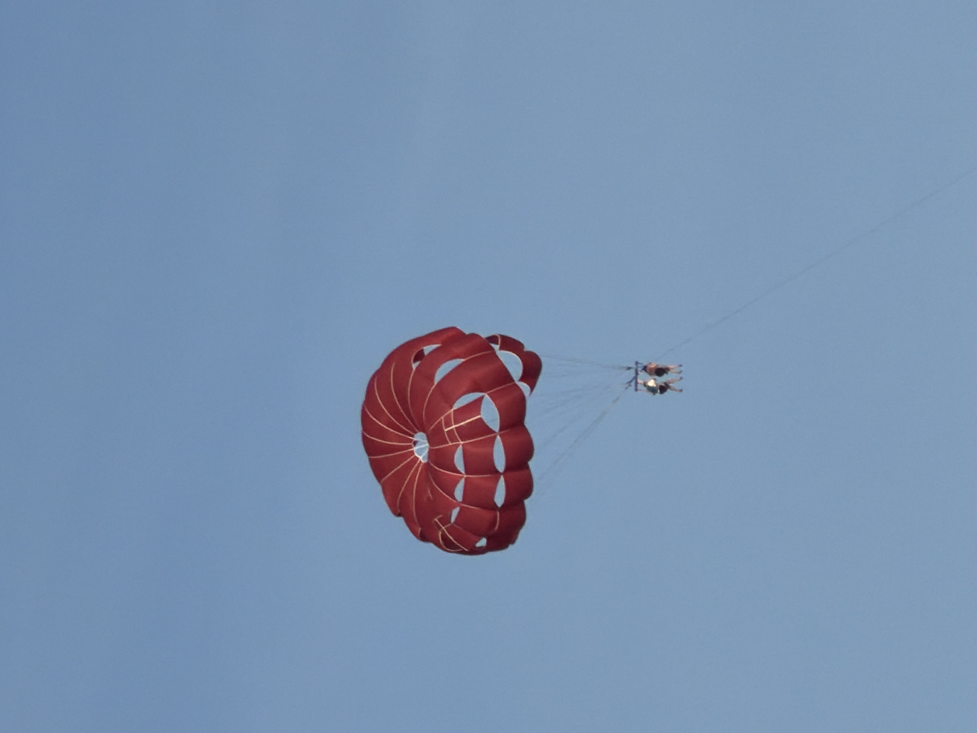 Two people are seen parasailing high in a clear, blue sky, presumably over the coastal area of Budva, Montenegro. The primary subject is a vibrant red and white parasail canopy, which is fully inflated and appears to be in mid-flight. The canopy is intricately designed with visible seams and structural lines, showcasing its rounded, open shape.
Suspended directly beneath the parasail are two individuals, linked by a network of ropes and harnesses to the main canopy. From this distance, their faces and specific attire are not clearly discernible, but they appear to be seated or hanging comfortably in harnesses, facing forward relative to the direction of travel (implied to be pulled by an unseen boat). A thin towline extends diagonally downwards from the parasailers, indicating their connection to a boat below the frame of the image.
The setting is characterized by a uniformly clear, light blue sky, suggesting bright daytime conditions and excellent weather for outdoor activities. There are no clouds or other atmospheric features visible, emphasizing the vast openness of the sky. The perspective is from below, looking up at the parasailers, making them appear high above. No other geographical features or objects are visible in the frame. There is no visible text.