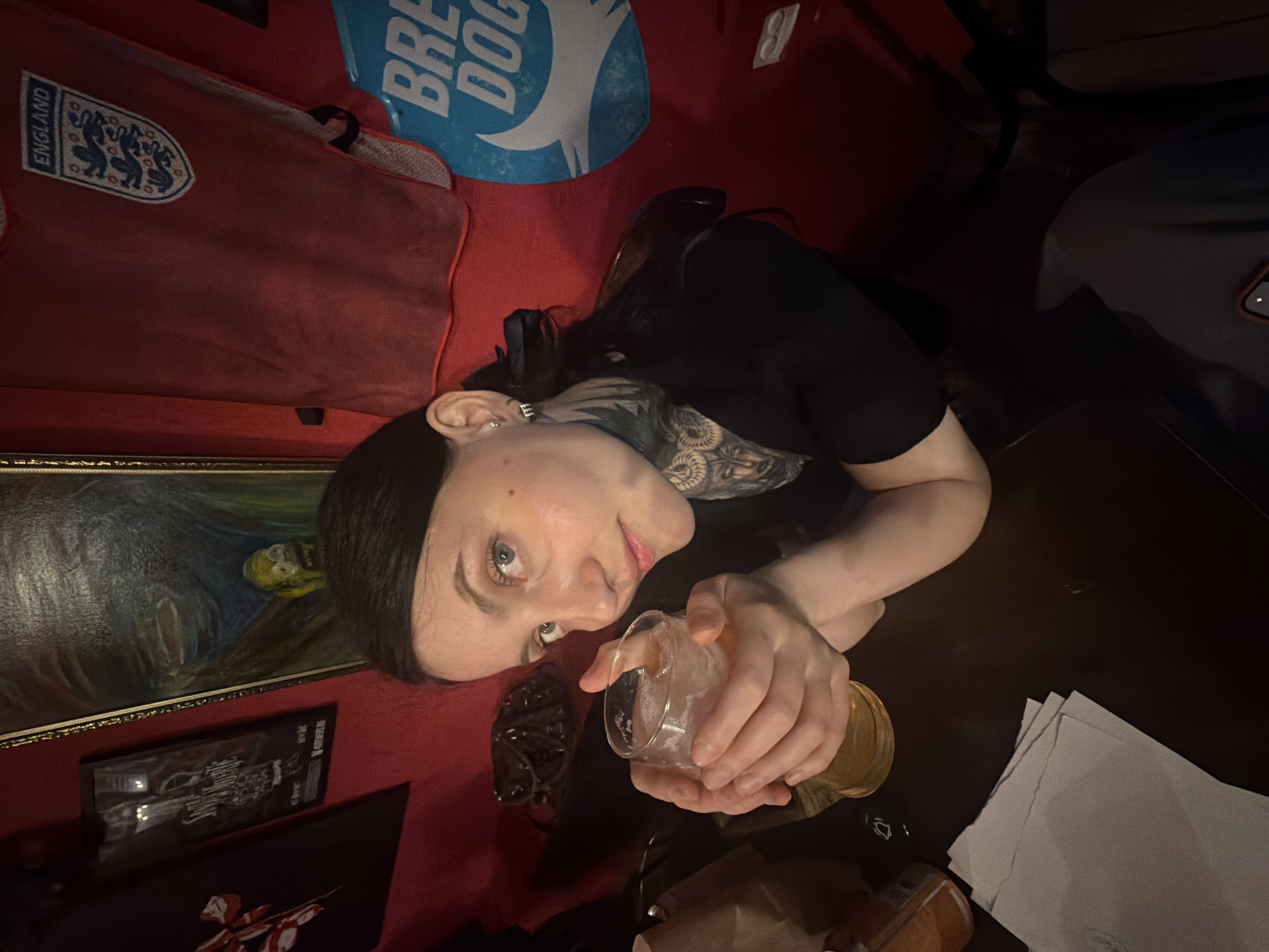 A woman is holding a glass of beer in an indoor setting. She has dark hair, light skin, and a tattoo on her neck. She is wearing a dark-colored shirt. The table is brown, with papers and a brown object. Behind her is a red wall. Also visible are an England flag and a BrewDog sign. I cannot determine if this image was taken in the city of Chaikovsky, Russia.