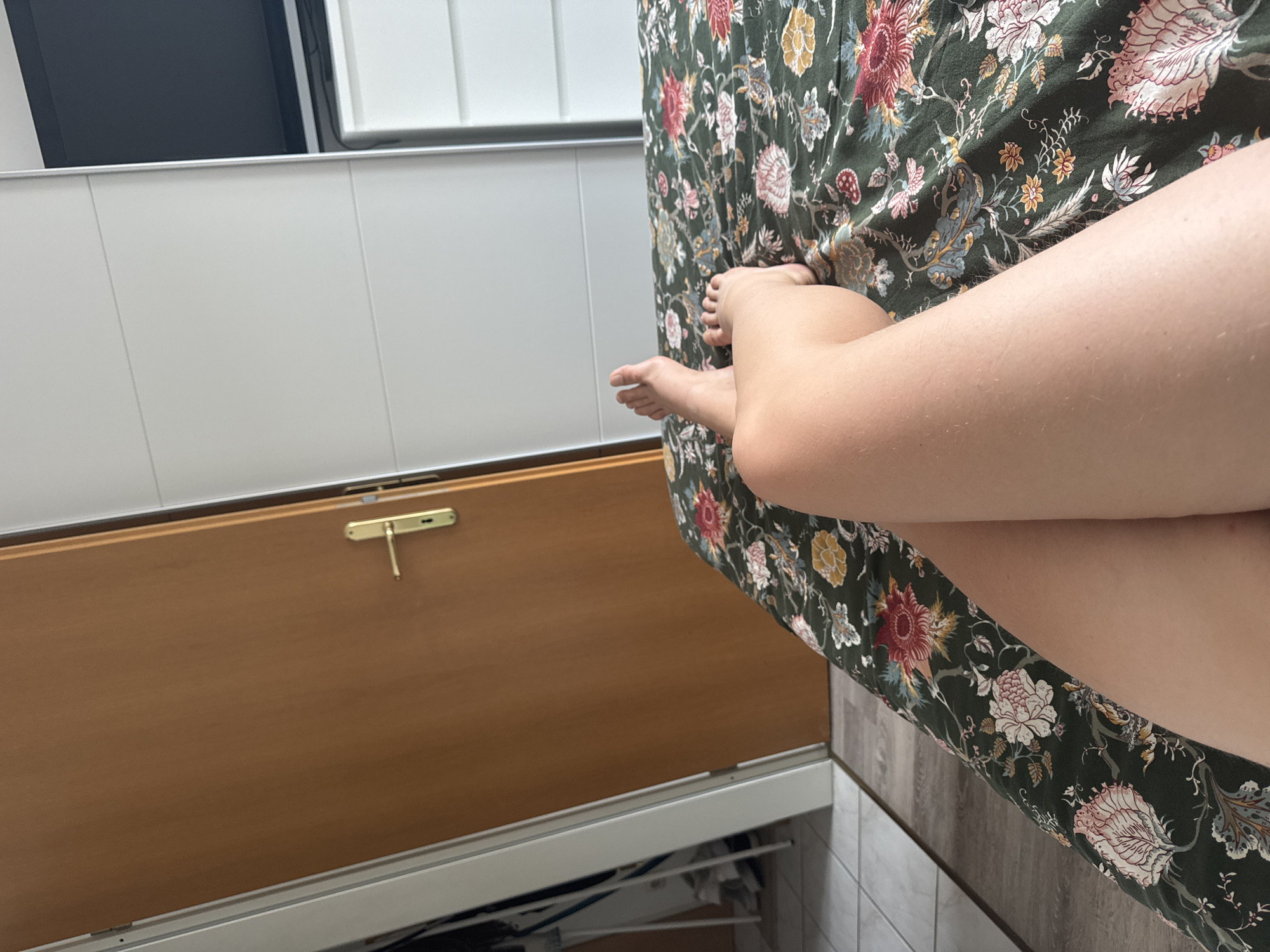 A close-up, point-of-view shot shows a person's legs crossed while lying on a bed. The bed is covered with a dark green floral-patterned comforter. In the background, to the left, is a closed, light brown wooden door with a gold handle. To the right of the door, a white wardrobe with horizontal panels is visible. The scene is likely indoors, possibly in a bedroom in Höhenkirchen-Siegertsbrunn, Germany.