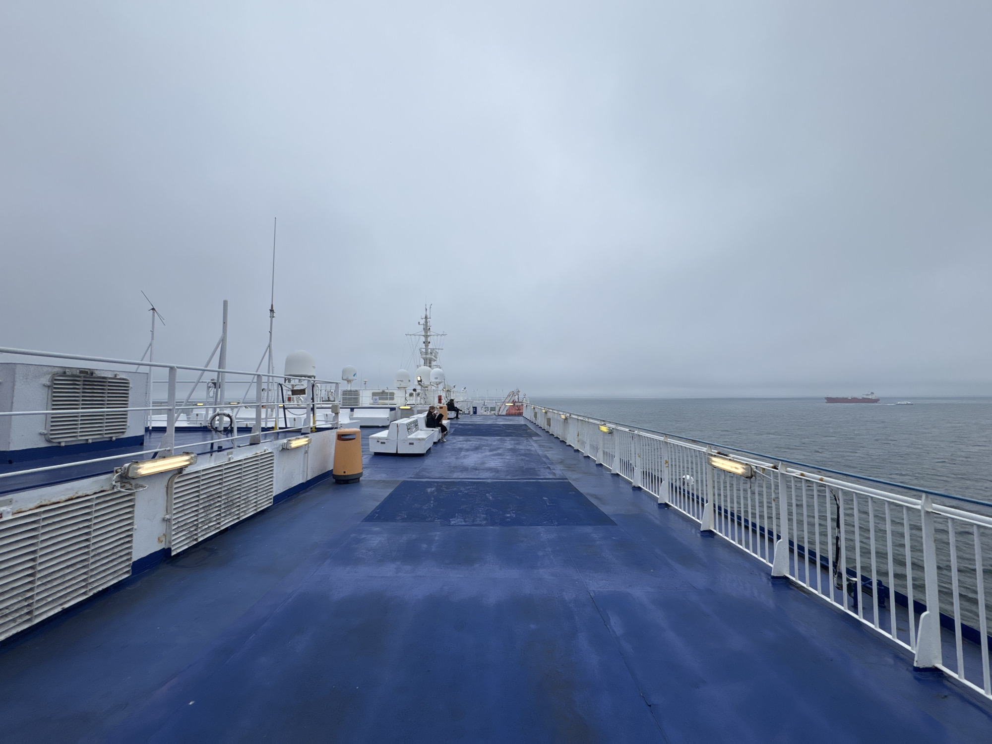 The image depicts a view from the deck of a ferry, likely on a cloudy day, with the sea and a distant cargo ship visible on the horizon. The deck itself is painted a vibrant blue, with white railings lining the perimeter. There are several pieces of equipment visible, including what appear to be air vents, lights, and communication antennas. In the middle ground, a few people are seated on white benches, and an orange trash receptacle is also present. The overall impression is that of a functional, utilitarian space on a large vessel. The image does not provide any specific visual cues to pinpoint its location in the city of Gdynia, Poland.