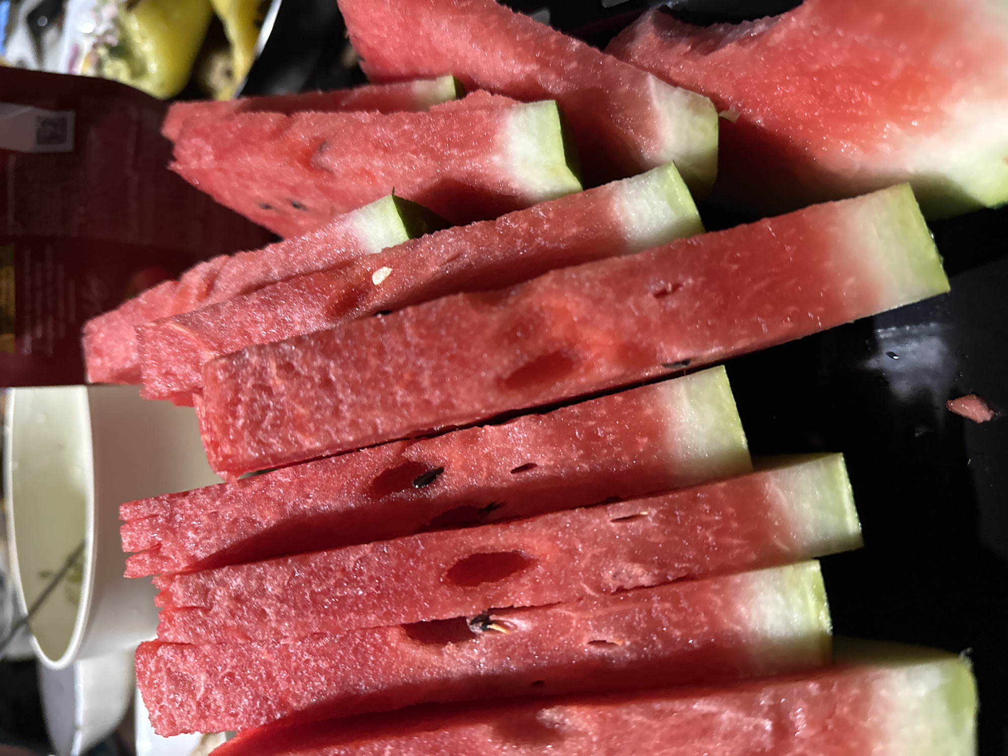 The image shows several slices of watermelon arranged on a black plate. The watermelon slices are cut into wedge shapes, with the red flesh and white rind visible. Some of the slices have black seeds. In the background, a white bowl with a ladle is visible, along with other blurred objects that suggest a domestic setting, possibly a meal or gathering. The lighting is artificial, and the focus is on the watermelon. There are no people visible in the image. The scene does not contain any specific information that would allow for the identification of the city of Sochi, Russia, or any particular location within it.