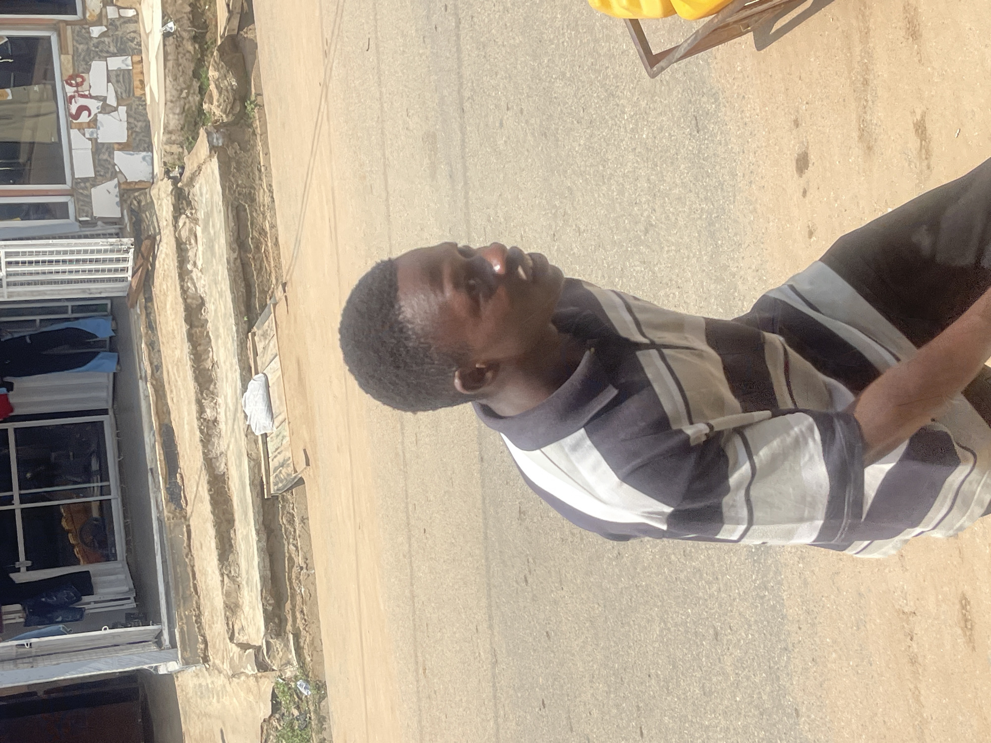 The image captures a street scene in Abuja, Nigeria, with a focus on a young man walking. He is wearing a dark blue and white striped polo shirt and dark trousers. The man is in the foreground, moving from left to right across the frame. Behind him, a section of a building with a cracked tiled facade is visible, featuring the partial inscription "576". Steps leading up to the building are present, with some debris on them. The ground is paved with asphalt and dirt. A small trolley containing yellow objects is partially visible to the right of the man. The lighting suggests it is daytime, with sunlight casting shadows.