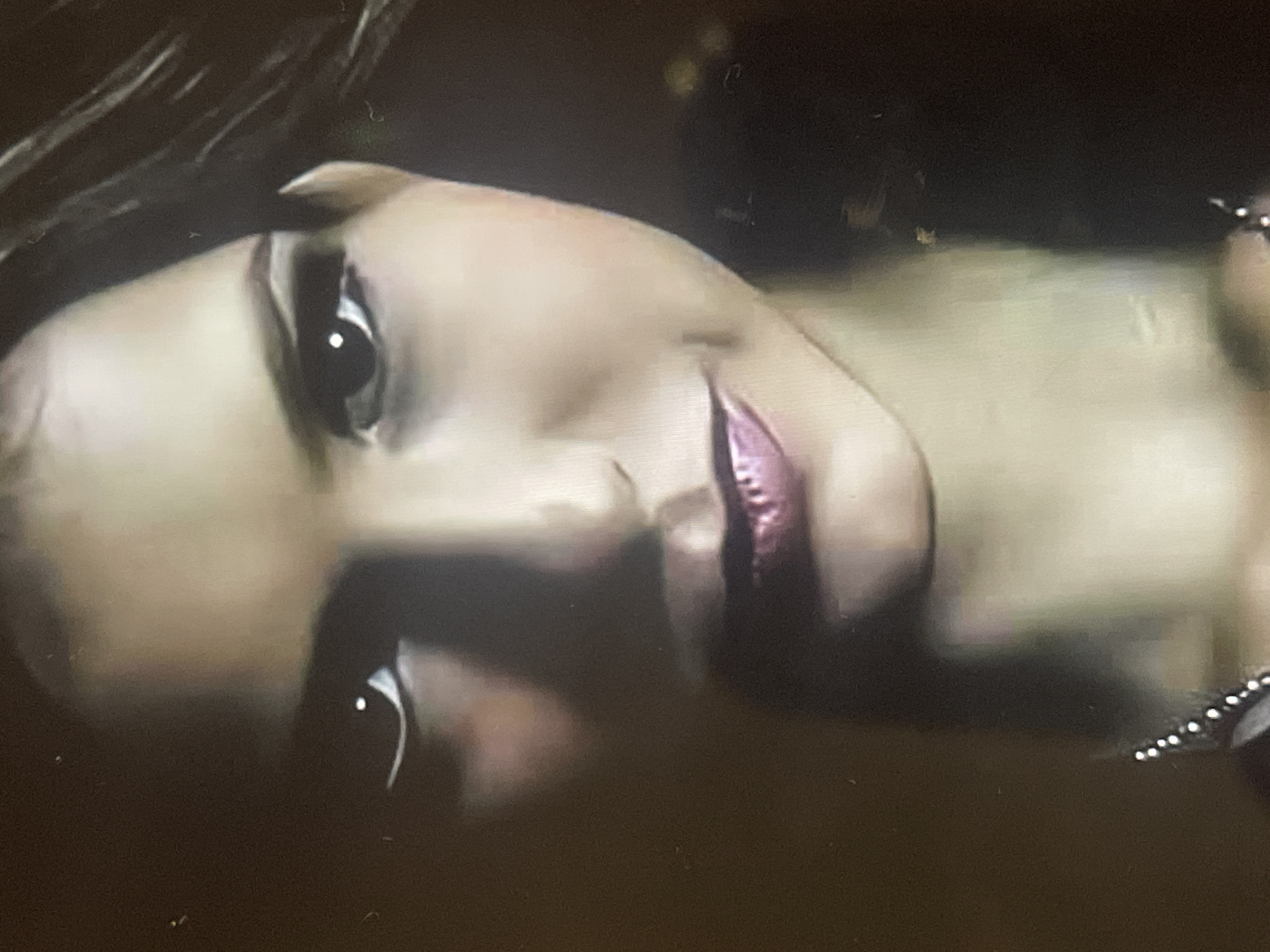 The image displays a close-up of a person's face, likely a woman, captured in what appears to be a low-light environment. The face is oriented horizontally in the frame, suggesting the person is lying down or the photo was taken from an unusual angle.
**Subjects:** The primary subject is an individual, with prominent features including wide-open eyes, a defined nose, and lips accented with shimmering pink or purple lipstick. Dark eyeliner or eyeshadow is visible around the eyes. Part of their hair, dark in color, can be seen in the upper left of the frame. On the right side, there's a glimpse of what appears to be a necklace or accessory adorned with small white beads.
**Scene and Setting:** The background is predominantly dark and out of focus, providing minimal environmental context. It suggests an indoor setting or nighttime conditions, with a few faint, indistinct light sources or reflections visible in the distance.
**Activity or Event:** The person's expression is somewhat ambiguous, characterized by wide, attentive eyes and slightly parted lips. They appear to be looking upwards or slightly to the side, potentially in a contemplative state, posing, or reacting to something off-camera. There is no clear activity depicted beyond the portrait-like pose.
**Notable Details:** The image quality exhibits some graininess and slight blur, indicative of a low-light capture or a still from a video. Catchlights are visible in the eyes, reflecting a light source. No visible text is present in the image. The provided context indicates this image is from Poltava, Ukraine.