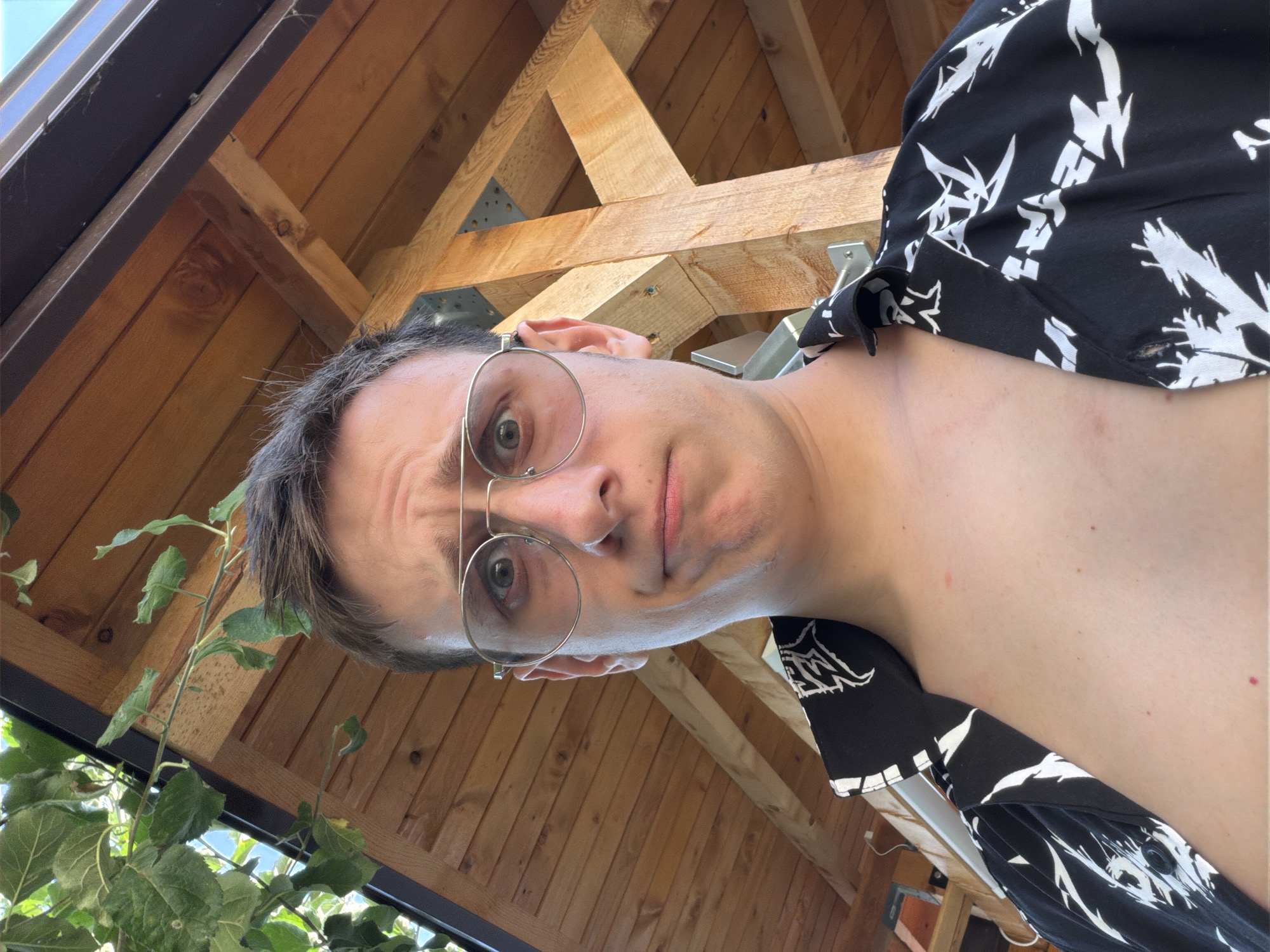 A close-up, low-angle selfie shows a young man with short, dark hair and a bewildered expression. He is wearing large, round, silver-rimmed glasses and a black short-sleeved shirt with a white abstract pattern. The background consists of a wooden structure, possibly part of an outdoor gazebo or patio cover, with visible wooden beams and planks. Green leaves from a tree hang from the upper left corner, suggesting an outdoor setting. The lighting indicates it is daytime. There are no other people or significant objects visible in the frame. The scene is likely in Bozhurishte, Bulgaria, given the context of the request.