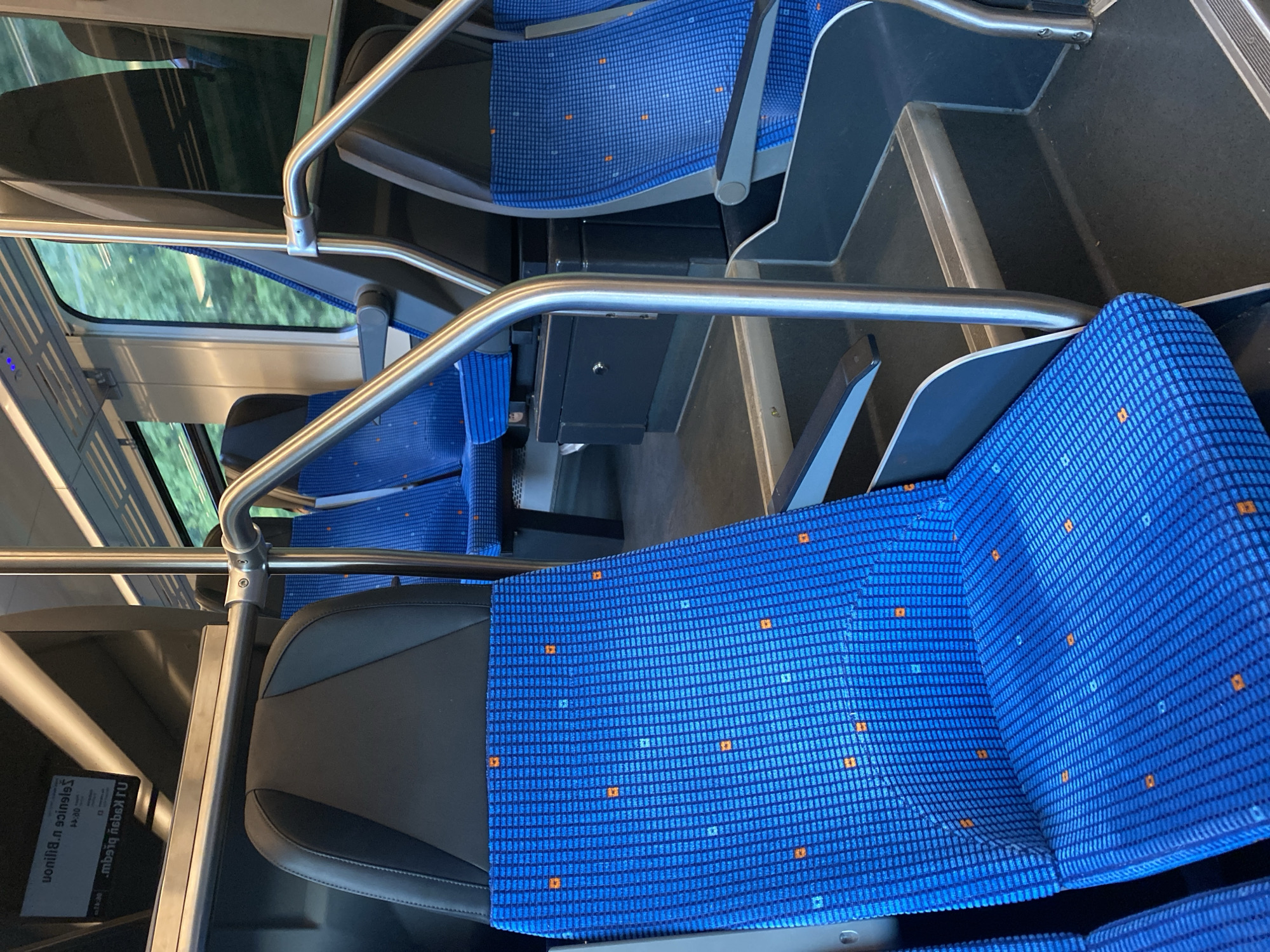 The image shows the interior of a train carriage, likely in the city of Bílina, Czechia, due to the displayed destination information. The focus is on the blue, patterned seats and metal handrails. Several empty seats are visible, upholstered in a bright blue fabric with a subtle checkered pattern and scattered orange squares. The seats are arranged in rows, with some appearing to be forward-facing and others backward-facing. Metal poles and handrails are strategically placed for passenger support, extending from the floor to the ceiling. In the background, through the windows, lush green trees and foliage are visible, suggesting the train is in transit through a natural landscape. A digital display screen mounted above the seats shows text in a foreign language, with "Bílina" and "Ústí n. L." (Ústí nad Labem) being partially discernible. The overall scene depicts an unoccupied public transport vehicle, emphasizing its functional design and clean, modern aesthetic.