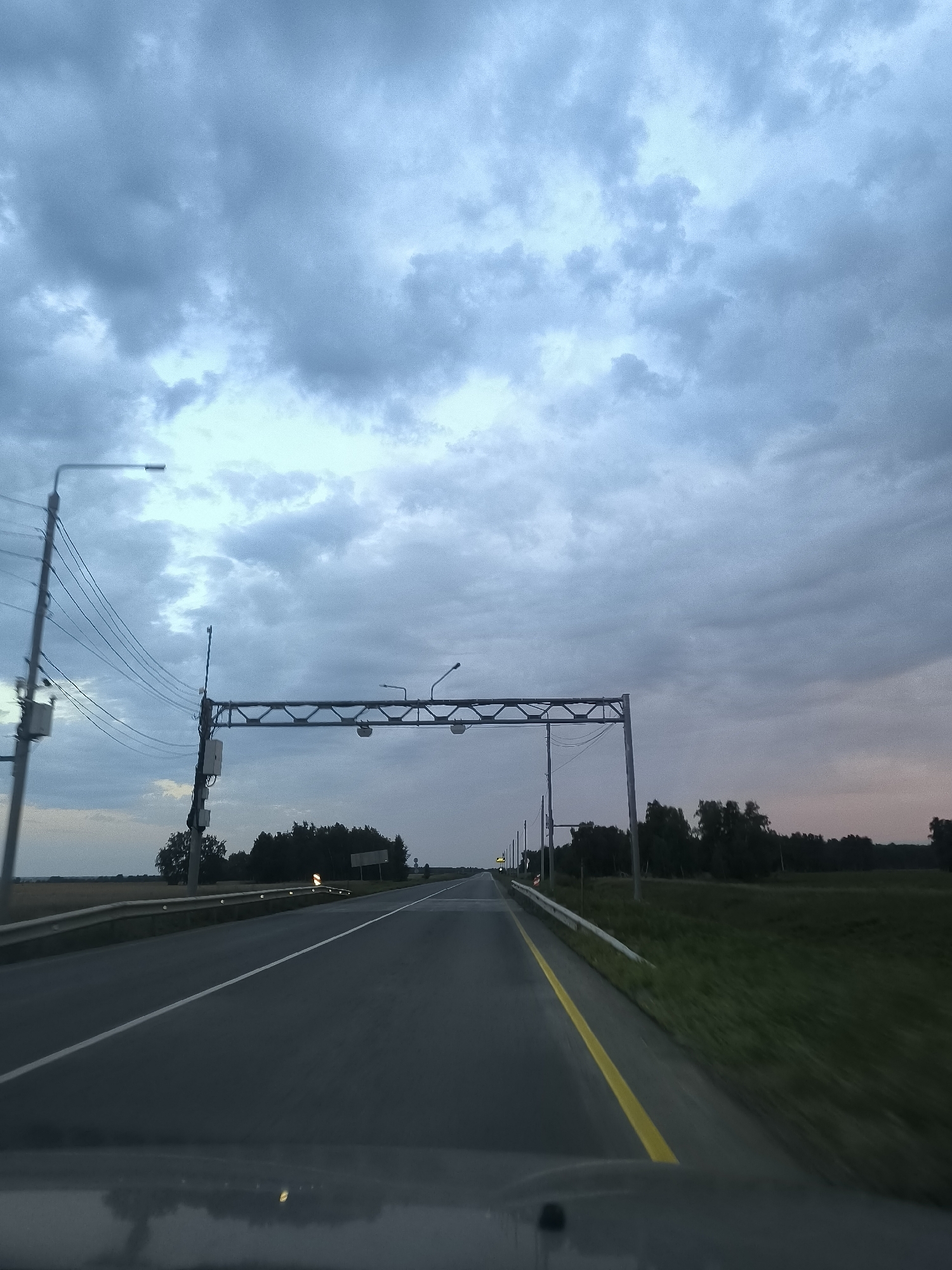 The image shows a view from a vehicle driving on a road in Avly, Russia. The sky is cloudy and a dusk or dawn light is present. There are power lines and streetlights along the side of the road. A metal overpass with lights spans the road in the distance. Trees and fields are visible on either side of the road. The road has yellow and white lane markings. The foreground shows the dashboard of the vehicle.