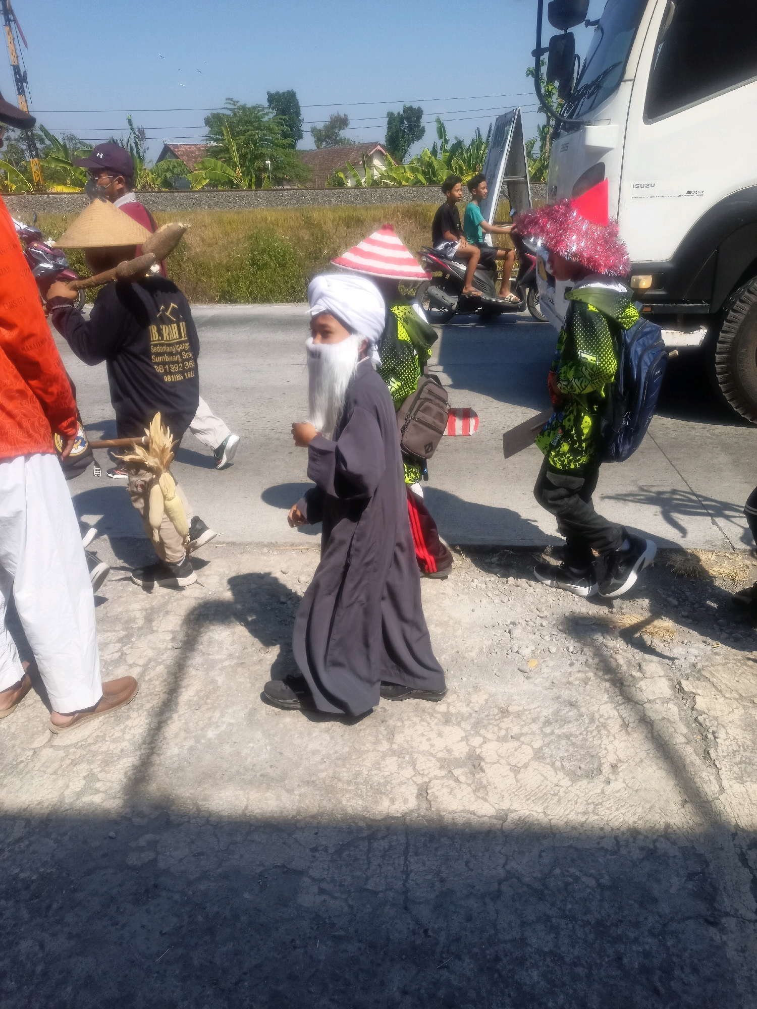 In the city of Sumberlawang, Indonesia, a group of individuals, primarily children, are participating in a parade or procession. The foreground is dominated by a child dressed in a dark robe and a white turban, with a fake white beard, suggesting a costume or religious attire. To the left, another child is walking, carrying a wooden pole over their shoulder with a straw hat on their head. Partially visible to the left is a person in an orange shirt and white pants. In the background, two boys are seated on a motorcycle, facing towards the viewer. A white Isuzu truck is parked on the right side of the frame, its front partially visible. The environment appears to be a roadside with a cracked asphalt surface and sparse vegetation. The sky is clear and blue, indicating a sunny day. The overall scene suggests a community event or celebration.
