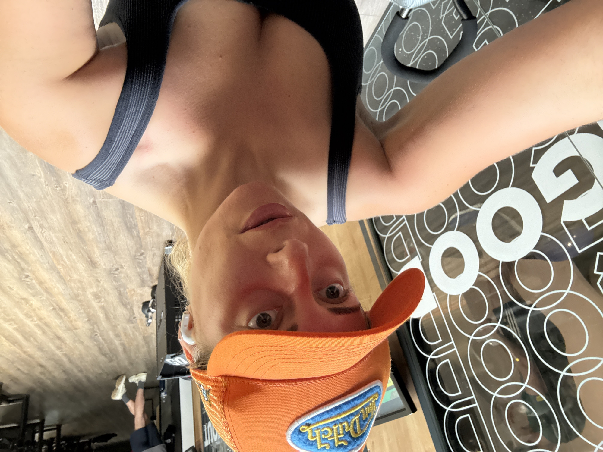 A woman with fair skin and blonde hair is taking a selfie indoors, likely in a gym. She is wearing a black sports bra and an orange Von Dutch cap. Her facial expression is neutral, and she appears to be looking directly at the camera. In the background, a person can be seen lying on the floor, presumably working out. There is exercise equipment visible, including what seems to be a stationary bike or similar machine with circular patterns on its surface. The floor is wooden, and gym equipment is visible in the background. Given the "GOOD OUT" logo on the equipment mat, the location is likely related to a gym or fitness studio. I can not verify a location within Paris, France.