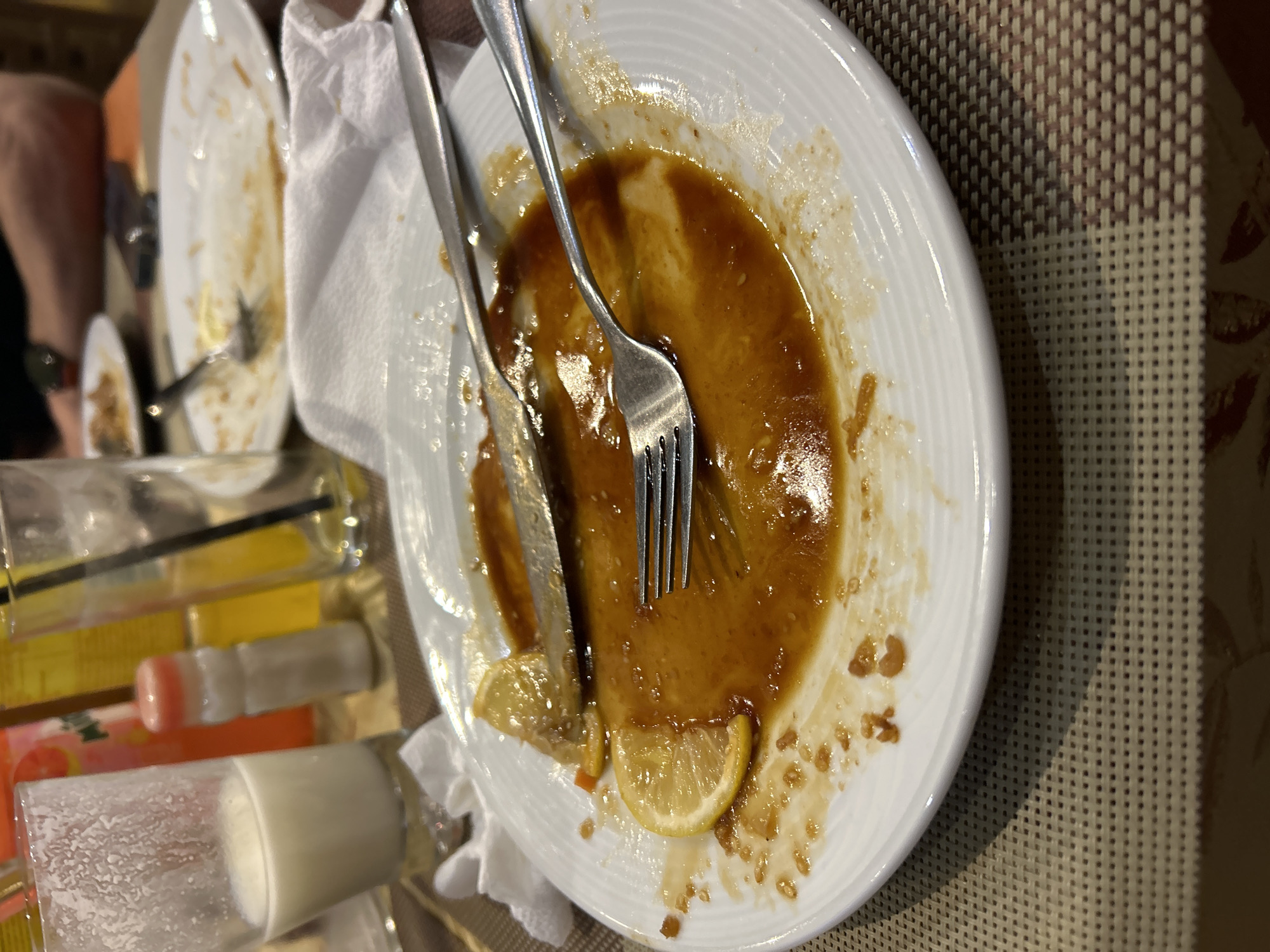 The image shows a dirty white plate with leftover brown sauce, a fork, and a knife on top of a white napkin. There are two lemon slices on the plate. Other dirty plates are partially visible in the background along with drinks. The location is Varna, Bulgaria. The environment is a dining table with a beige woven placemat. A person's arm is visible on the left side of the frame.