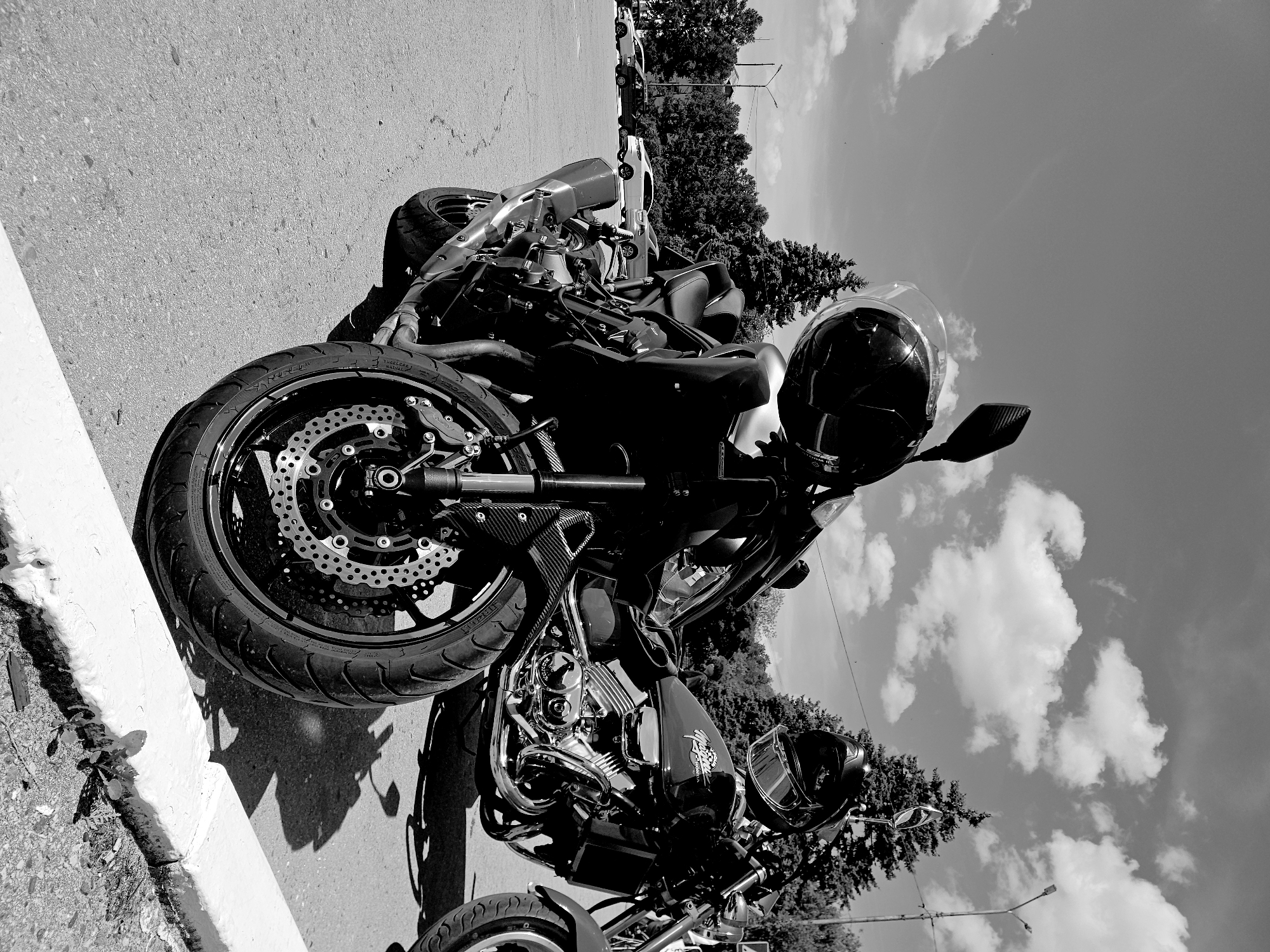 The image depicts two motorcycles parked on an asphalt surface adjacent to a white curb. A black helmet with a clear visor is positioned on the handlebars of the motorcycle in the foreground. The front wheel and part of the engine of this motorcycle are clearly visible. Behind it, a second motorcycle, a Honda, is partially visible, also with a helmet on its handlebars. In the background, trees and parked cars line a street, suggesting an urban or suburban setting. The sky is bright with scattered clouds. The scene is rendered in black and white. The motorcycles appear to be parked in a lot or on a street in Ulyanovsk, Russia. No people are directly interacting with the motorcycles.