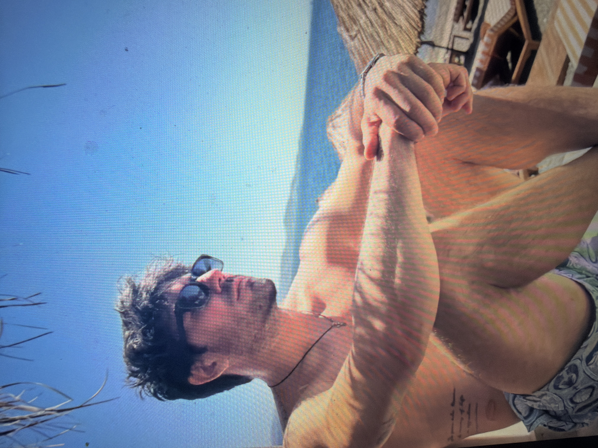 The image displays an adult male seated in a sunlit coastal environment. The subject is depicted from the waist up, facing towards the right side of the frame in profile. He is shirtless, wearing dark sunglasses, and has dark, wavy hair and a short beard. A thin black cord necklace with a small, indistinct pendant is visible around his neck. His arms are bent, with hands clasped together and resting on his bent knees. A tattoo with several lines of text is visible on his left rib cage area. He is wearing patterned shorts, primarily blue and white.
The background features a clear blue sky, transitioning to a calm blue sea with a discernible horizon line, and faint distant landmasses. To the upper left, dried plant material, possibly part of a thatched roof or palm fronds, overhangs into the frame. On the right side of the image, part of a thatched umbrella and several wooden lounge chairs with striped cushions are visible on a beach or wooden deck. The scene is brightly illuminated by direct sunlight. The image exhibits a prominent pixel grid, indicating it is a photograph taken of a digital screen. According to the provided information, the location is Shumen, Bulgaria.