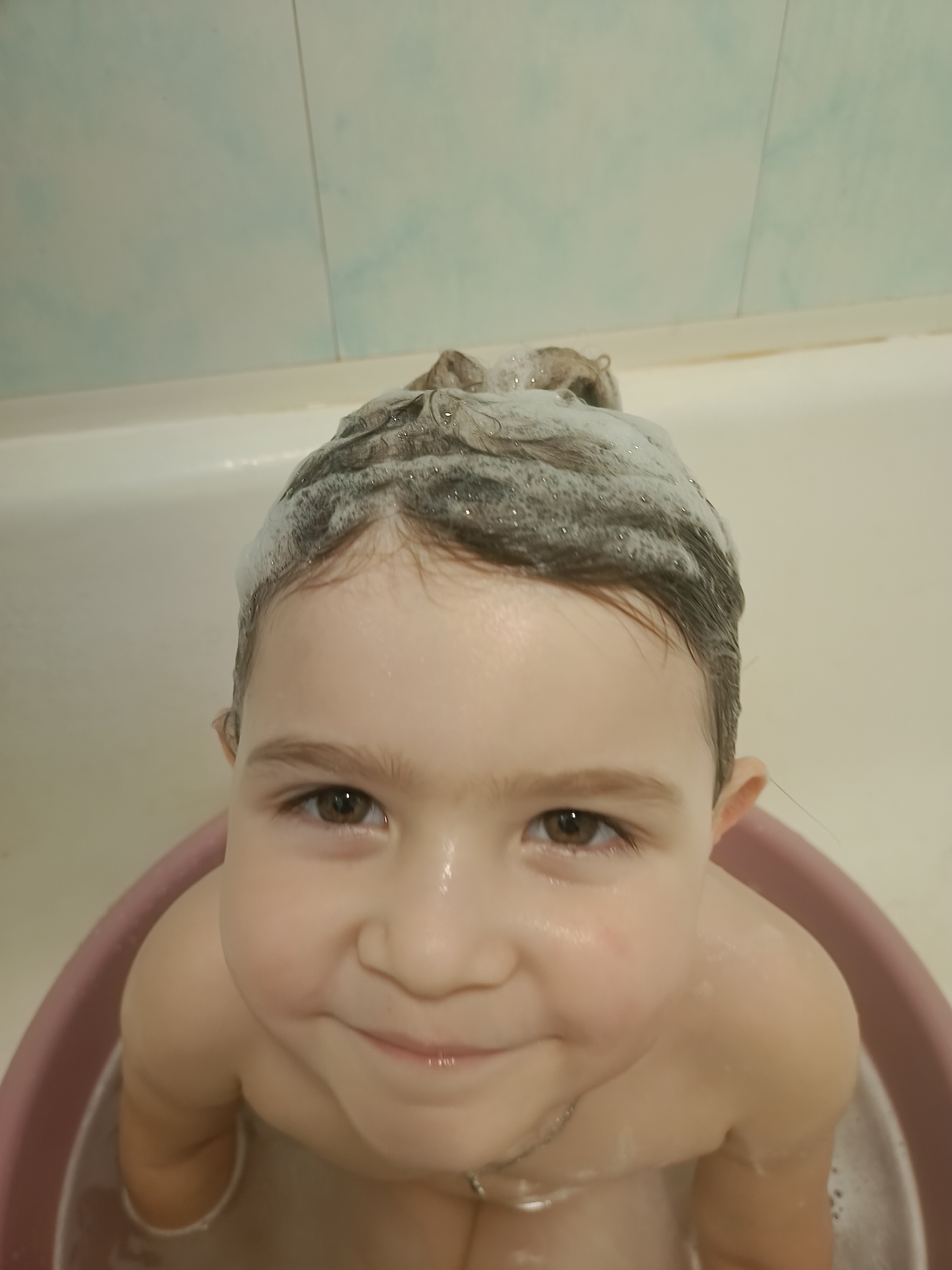 A close-up, high-angle shot shows a young child in a bathtub. The child has brown hair with shampoo lather on top of their head. Their eyes are a warm brown, and they have a wide, happy smile. The child is sitting in a pink bathtub, and some water is visible at the bottom. The background shows light blue tiled walls of the bathroom. The overall mood is joyful and playful, suggesting a bath time routine. The location is identified as Khokhly, Russia. There is no visible text in the image.