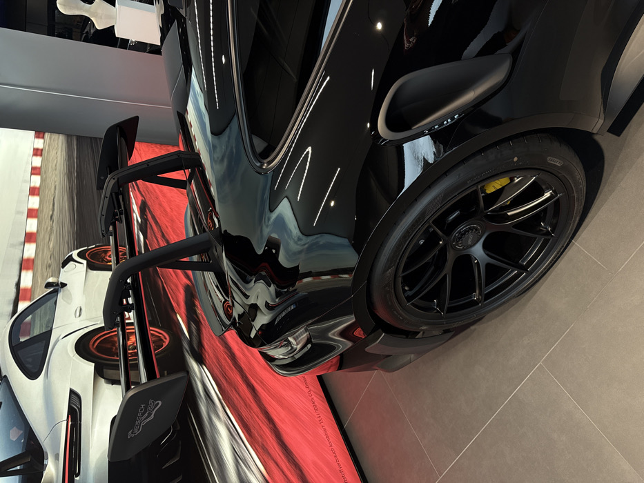 This image captures an indoor luxury sports car showroom in Regensburg, Germany. The primary subject is a highly polished gloss black Porsche 911 GT3 RS, viewed from its rear three-quarter angle. The vehicle exhibits a prominent black swan-neck rear wing, black multi-spoke center-lock wheels, and distinct yellow brake calipers visible through the spokes. Pirelli P Zero tires are mounted, with sidewall details observable. The car's reflective surface distorts and mirrors ceiling lights and other environmental elements.
In the background, a large digital display or banner spans the wall, featuring a white Porsche 911 GT3 RS with orange wheels and a "WEISSACH RS" branded rear wing, depicted on a racetrack with red and white curbing. German text regarding fuel consumption is partially visible at the bottom left of this display, stating "Kraftstoffverbrauch kombiniert: 13,4 l/100 km. CO₂-Emiss..." The showroom floor consists of light gray tiles with visible grout lines. A white abstract sculpture or decorative object on a white pedestal is partially visible in the far right background. The scene is static, with no individuals or active interactions present.