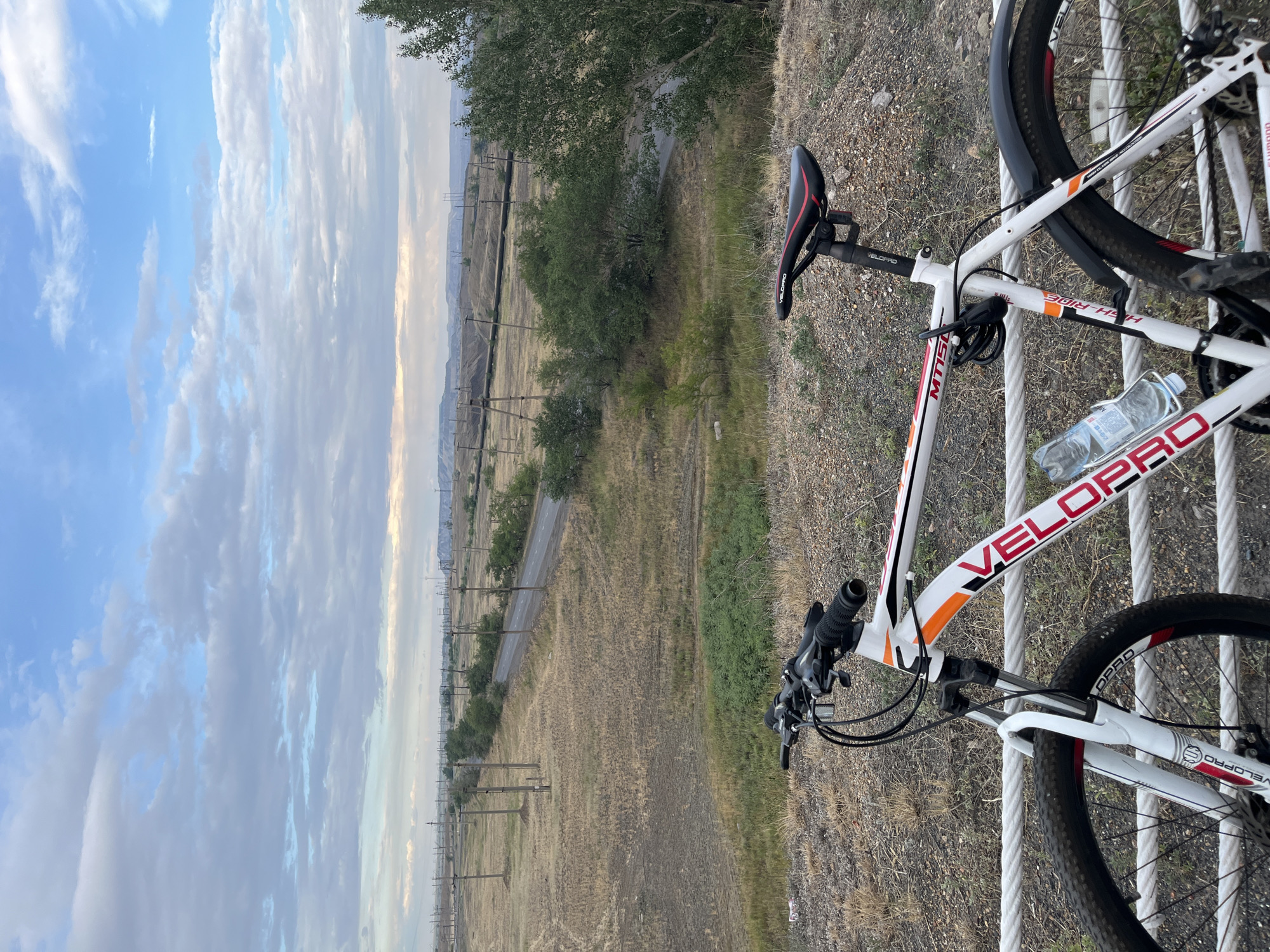 A white bicycle with orange and black accents is parked in the foreground, resting on a white rope railing. The bicycle has a "VELOPRO" logo on its frame, a black seat with red trim, and a water bottle attached to a cage. Behind the bicycle, a long, straight asphalt road recedes into the distance, flanked by dry, grassy fields with numerous utility poles. The landscape is somewhat industrial, with power lines and what appear to be mining spoil heaps or industrial structures visible in the background. The sky above is partly cloudy with patches of blue sky showing through. The overall setting suggests an outdoor location near Ekibastuz, Kazakhstan, likely a rural or semi-industrial area on the outskirts of the city, characterized by its open spaces and infrastructure related to power generation or mining. There are no people visible in the image.